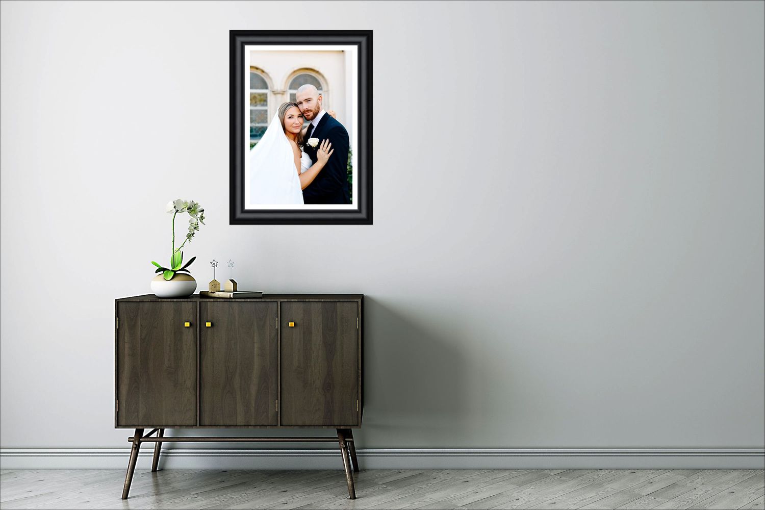 Framed wedding portrait of a couple embracing, displayed above a dark wood mid-century modern sideboard in a minimalist room.