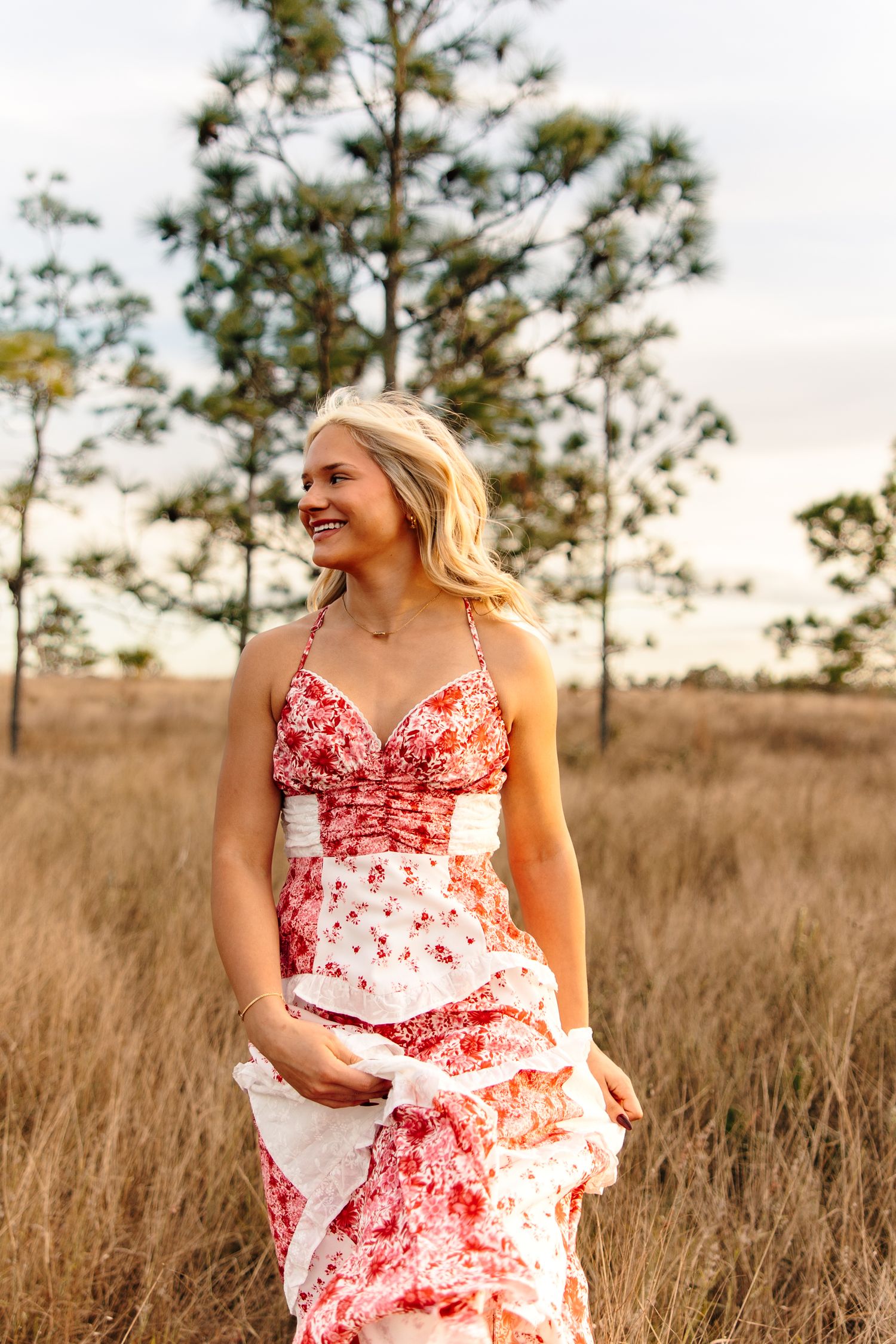 Smiling blonde woman in red floral maxi dress standing in golden grass field with trees in background.