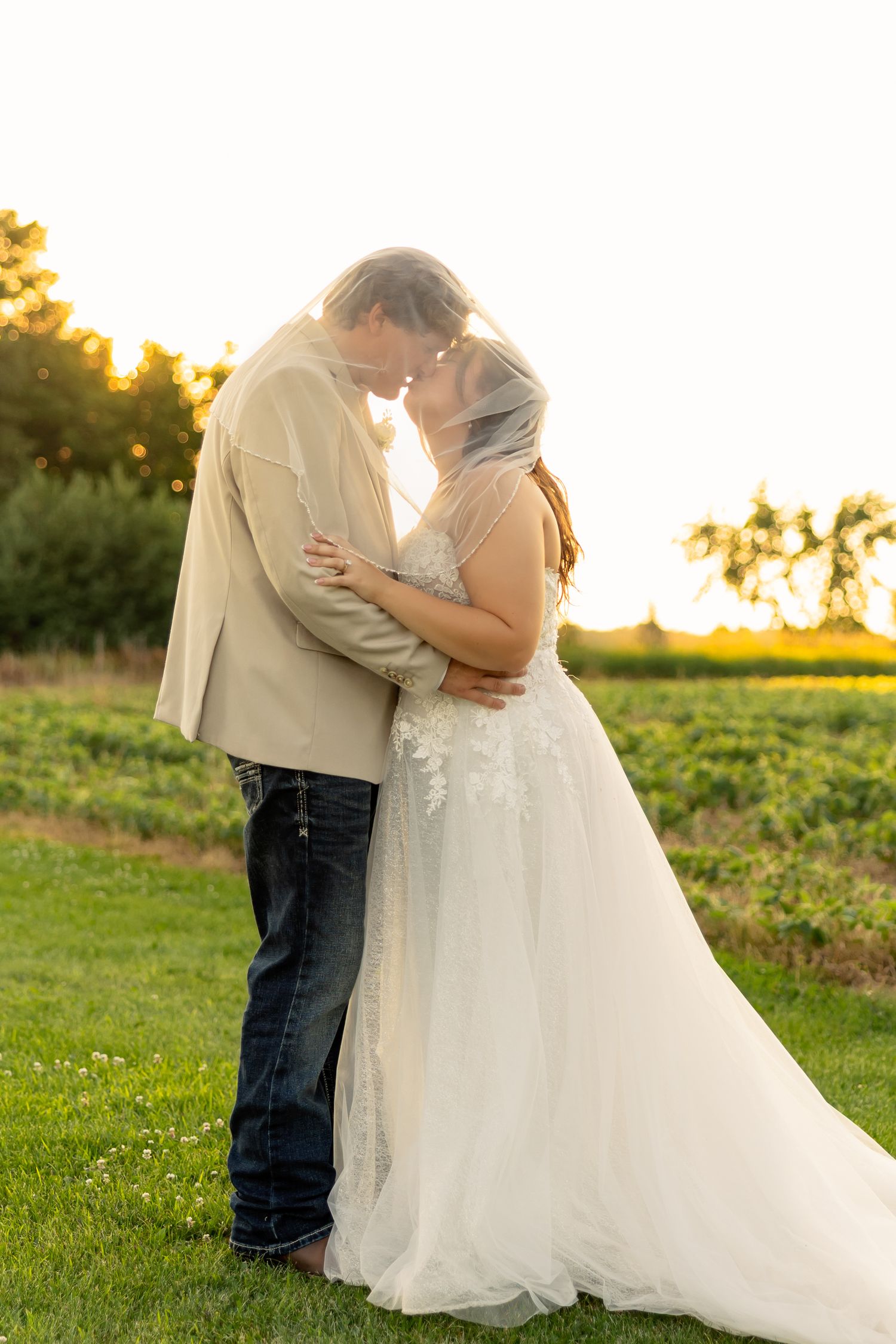 Bride and groom sharing a romantic kiss at sunset in a lush green field, bride wearing a white lace gown with veil.