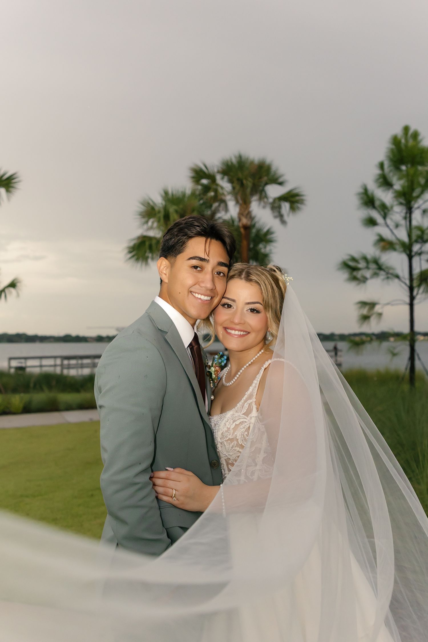 Bride and groom embracing outdoors near waterfront with palm trees, bride wearing lace gown and flowing veil.