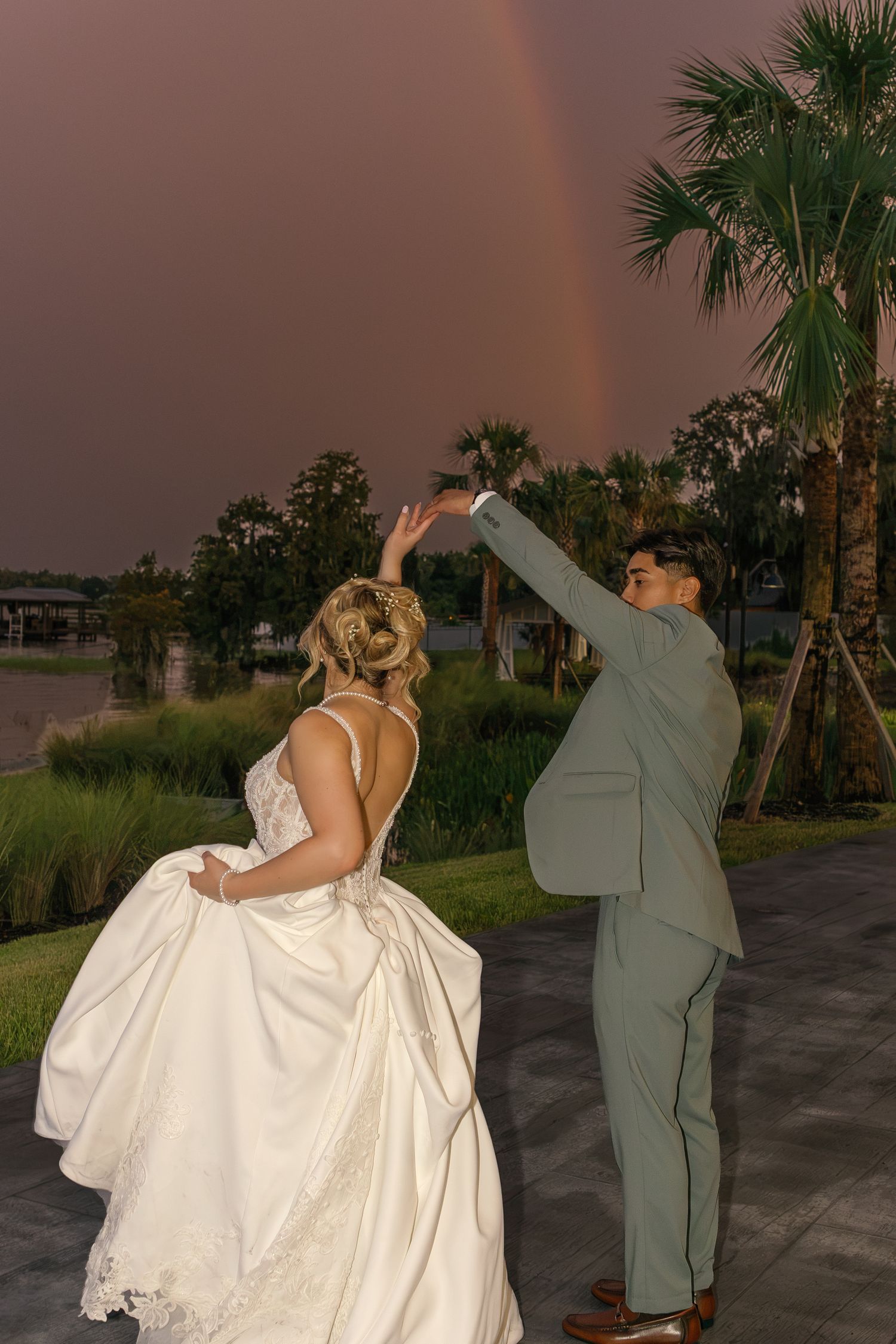 Bride and groom dancing outdoors at sunset with a rainbow over palm trees in the background.