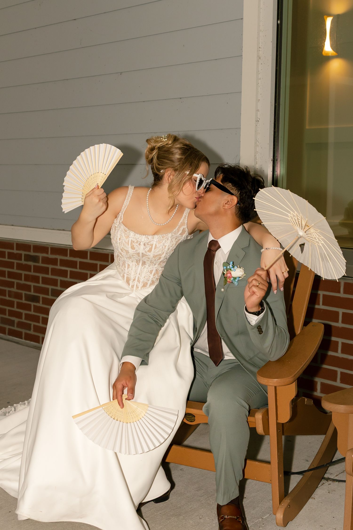 Bride and groom kissing while holding decorative fans and a parasol, wearing sunglasses in sage green and white wedding attire.