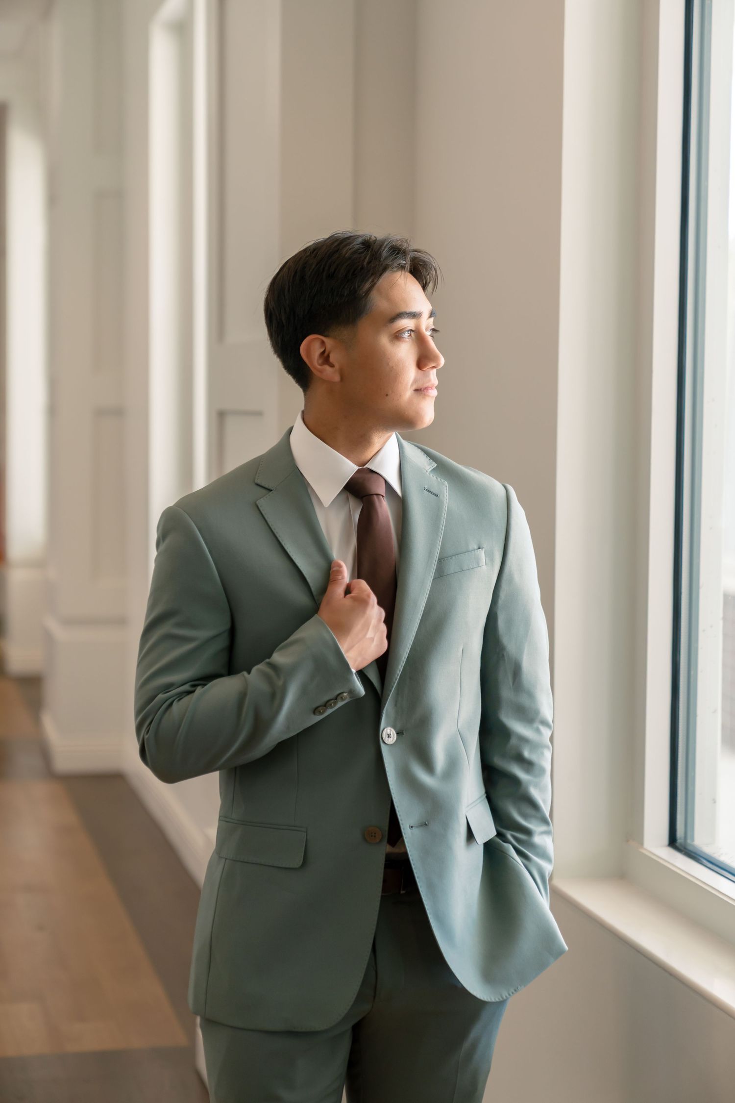 Young man in sage green suit and brown tie gazes out window, posing confidently in bright hallway.