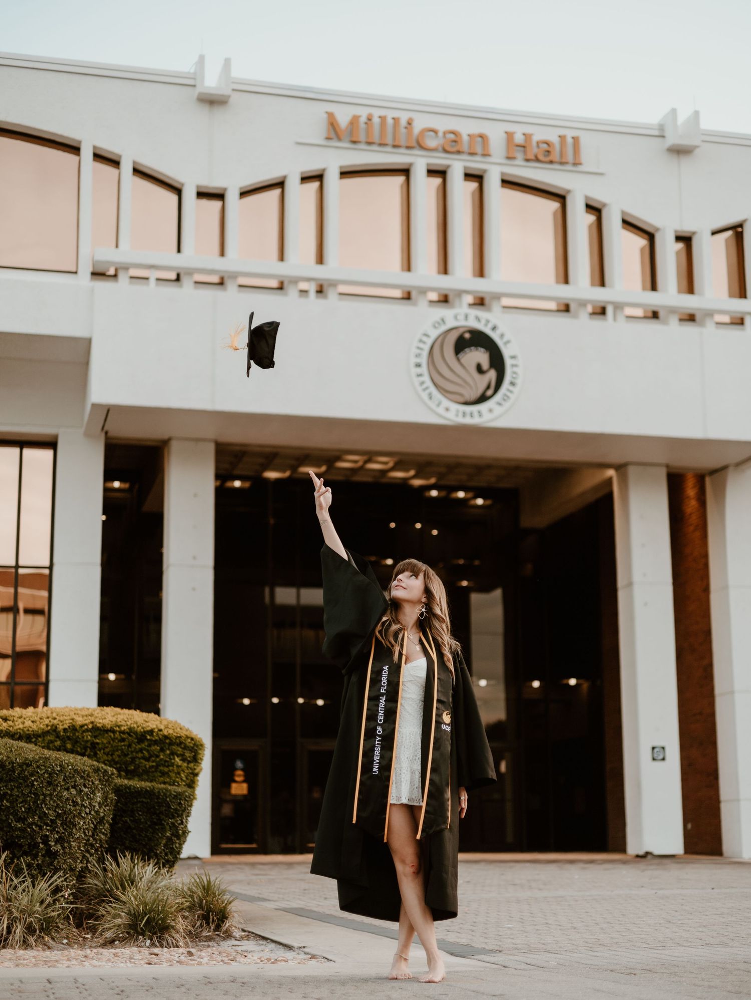 Graduate in cap and gown tosses mortarboard in front of Millican Hall at University of Central Florida.