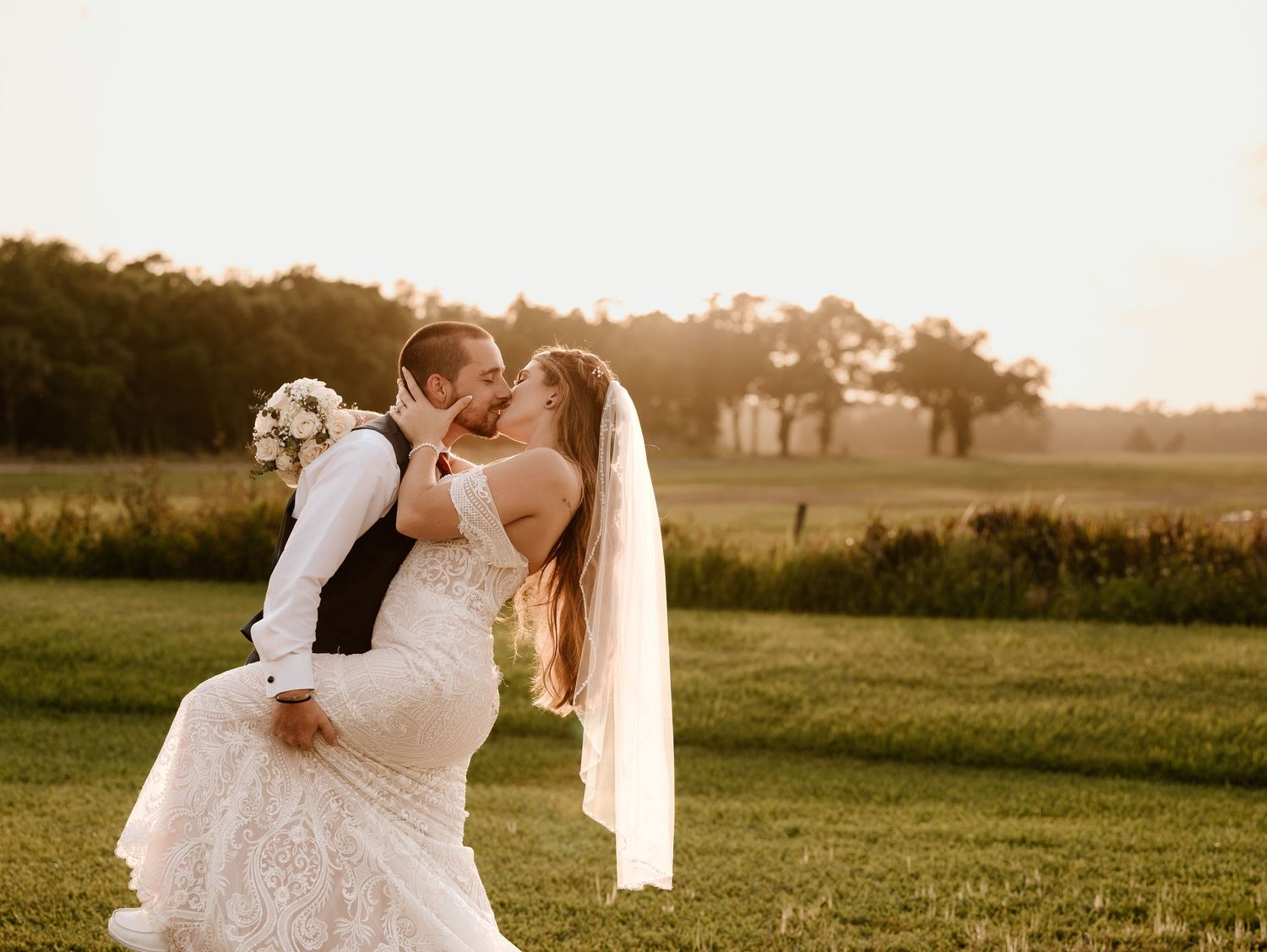 Groom lifting bride in wedding attire at golden hour on lush green field with trees in background.