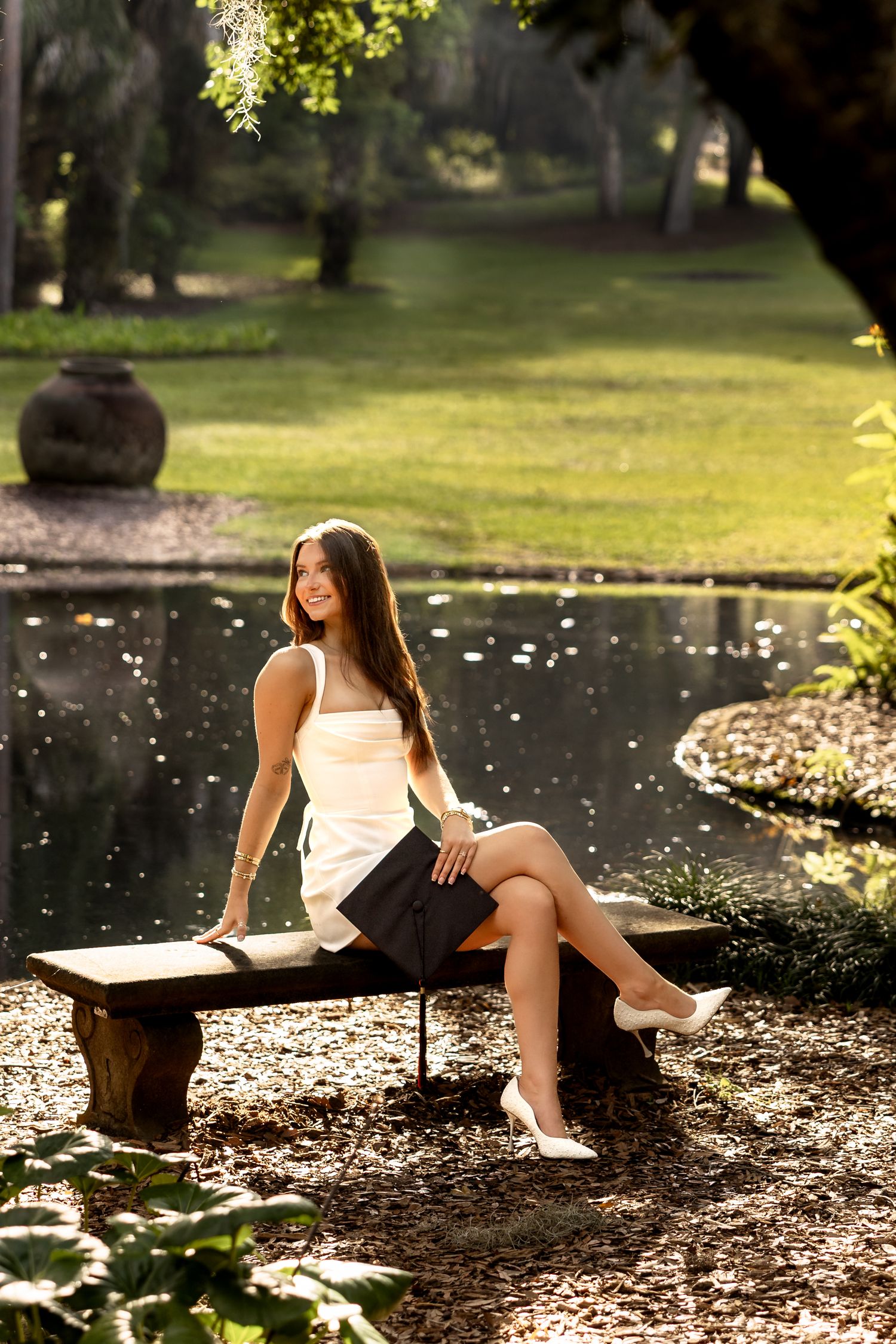 Young woman in white dress sitting on wooden bench by serene pond in lush green park, smiling in golden sunlight.