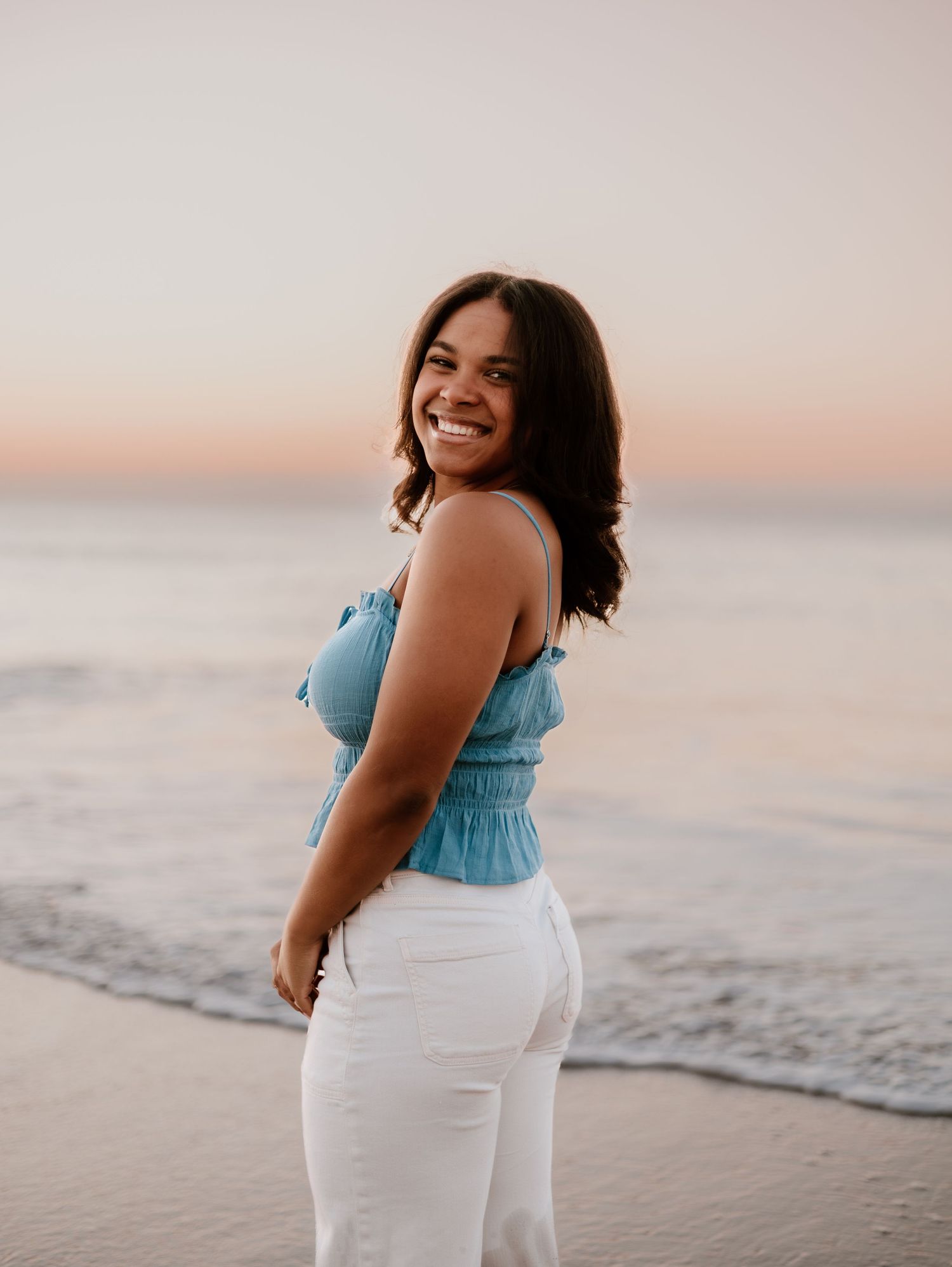 Person in blue top and white pants smiling on sandy beach at dusk.