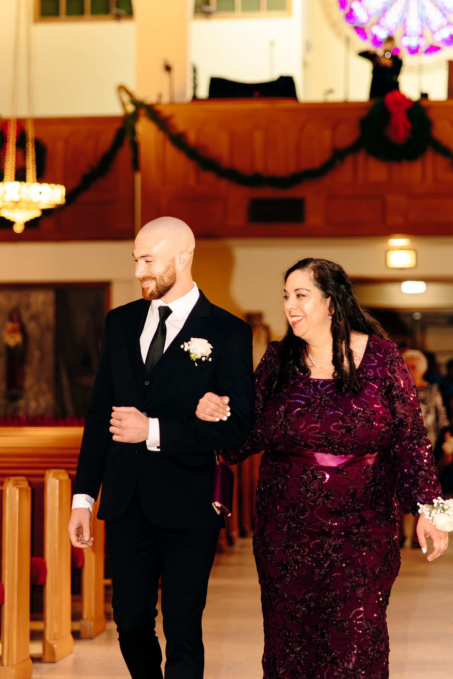 Groom in black suit escorts woman in purple sequin gown down church aisle during wedding ceremony.