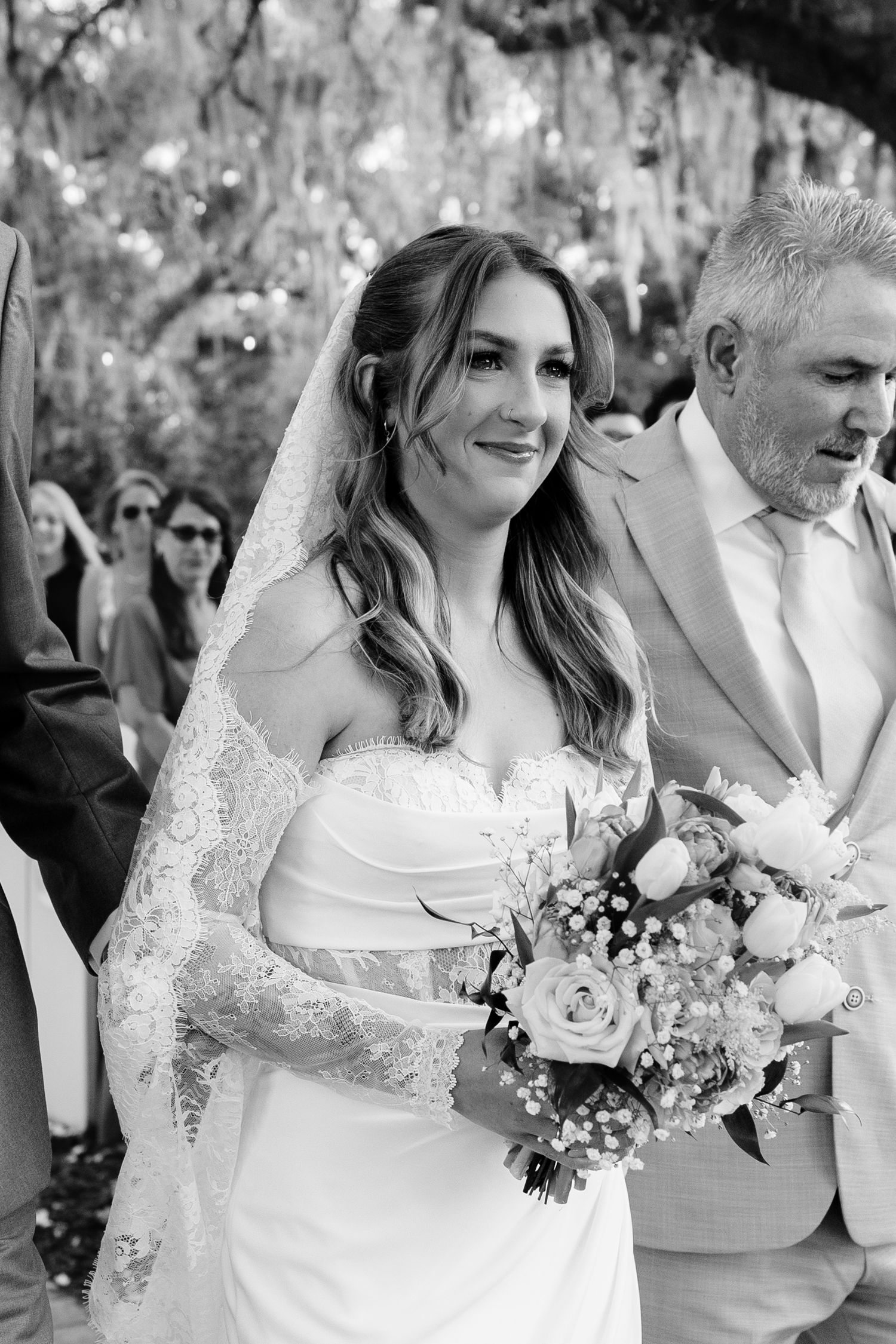 Black and white photo of a smiling bride in a strapless gown and lace veil holding a bouquet at an outdoor wedding ceremony.