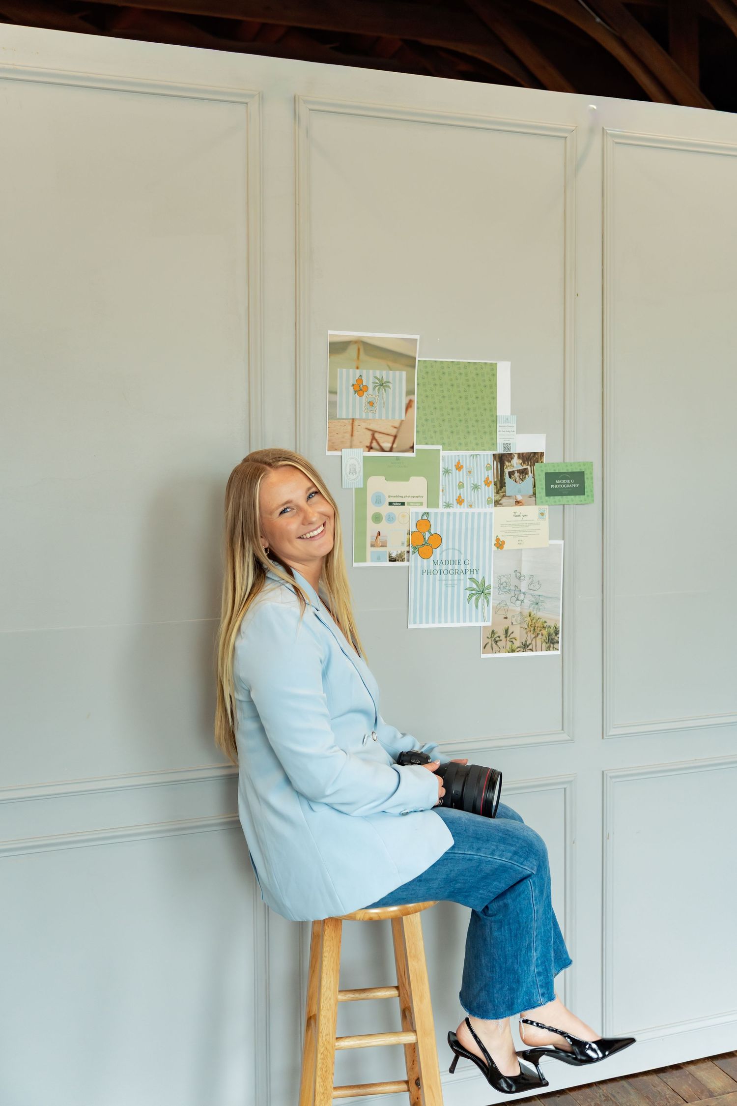 Smiling blonde woman in blue blazer and jeans sits on wooden stool near mood board with green and orange design samples.