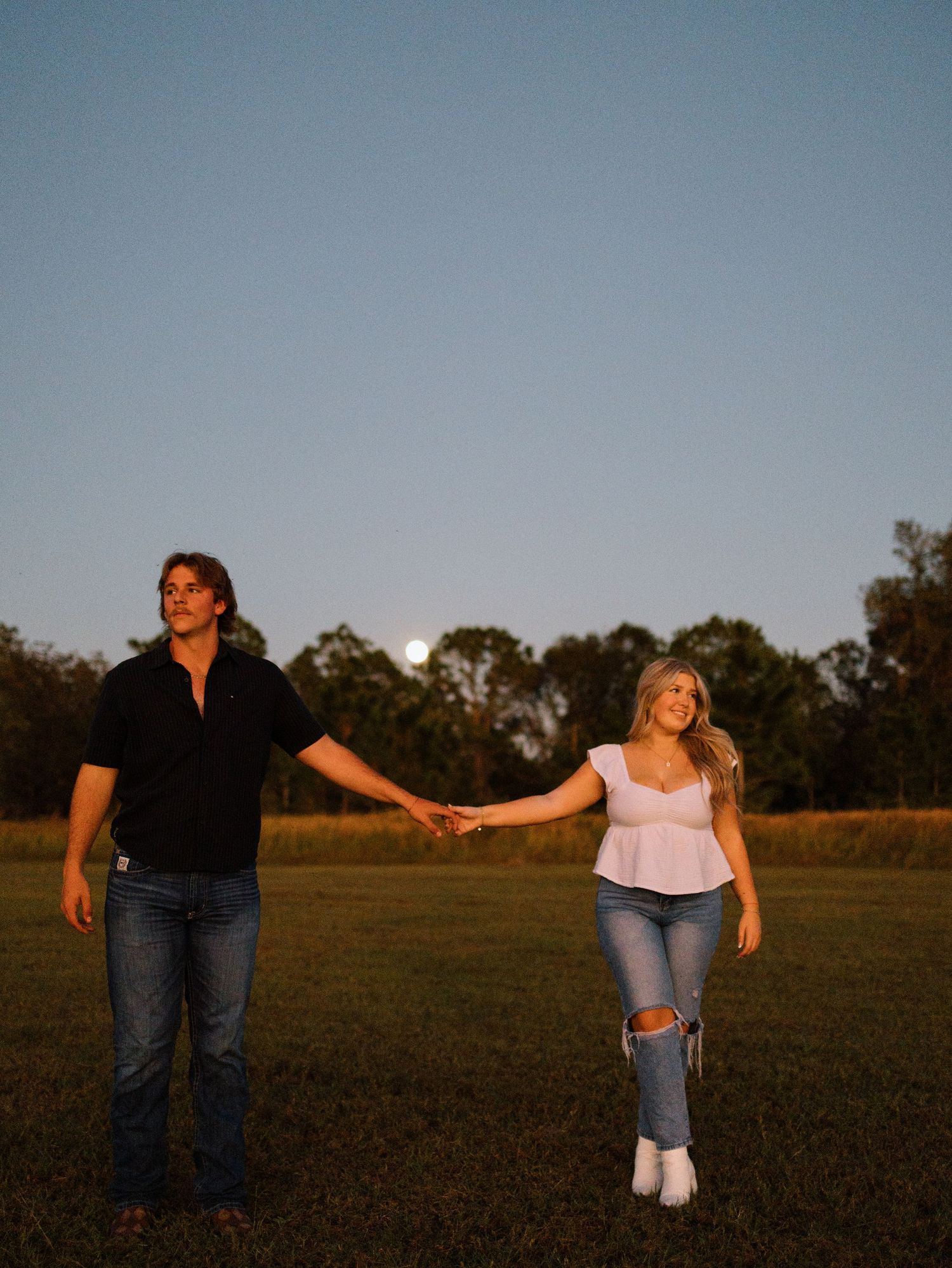 A couple holds hands while walking together in a grassy field at sunset with trees in the background.