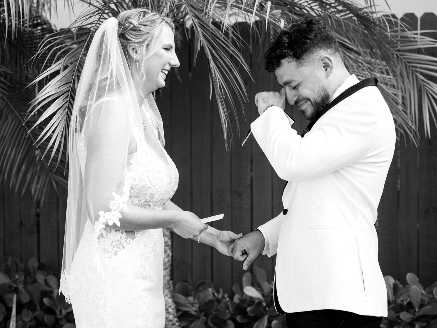 Black and white photo of emotional groom wiping tears as bride places wedding ring on his finger.