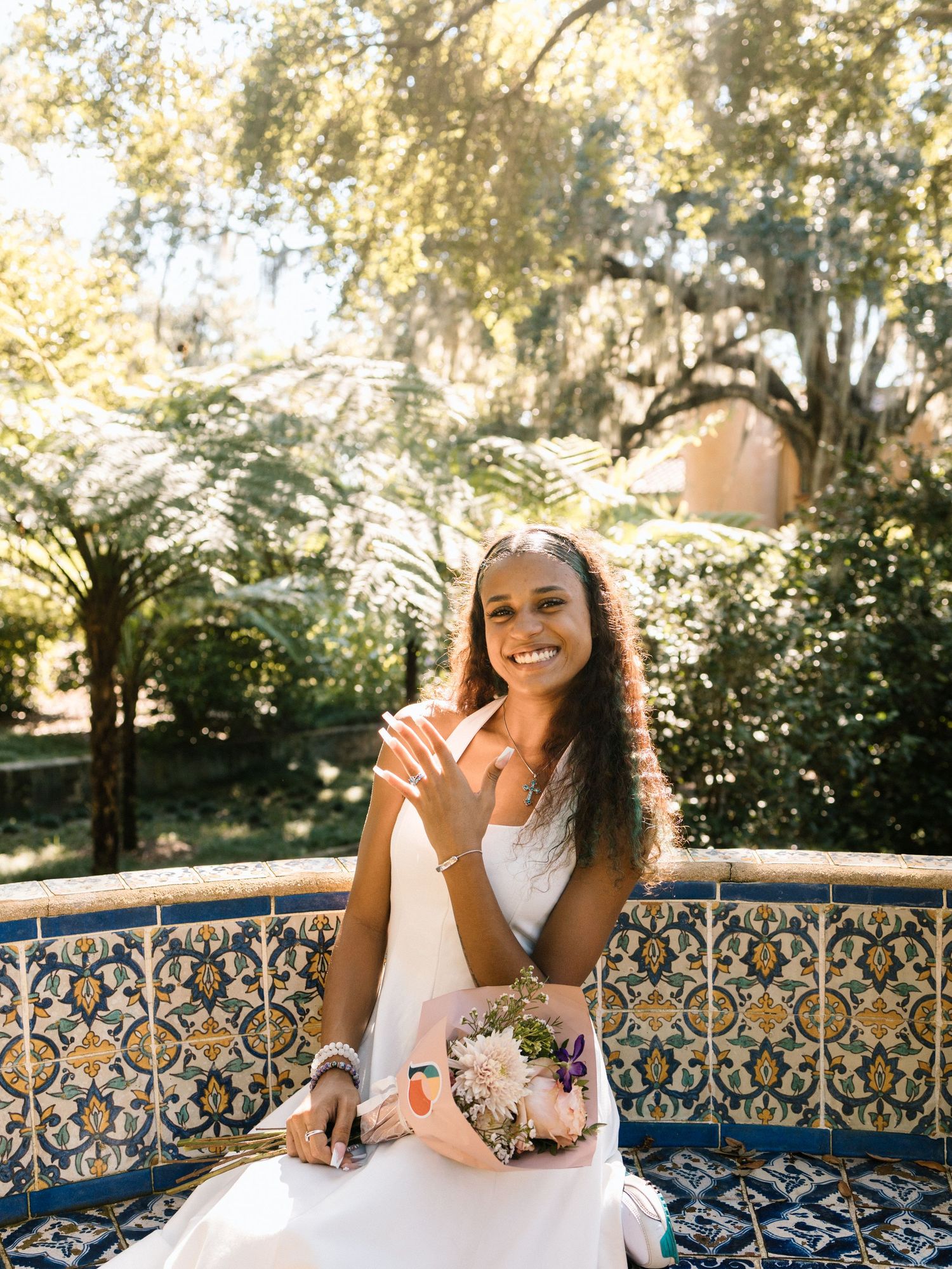 Woman with curly hair sits on decorative tiled bench in garden setting with natural sunlight filtering through trees.