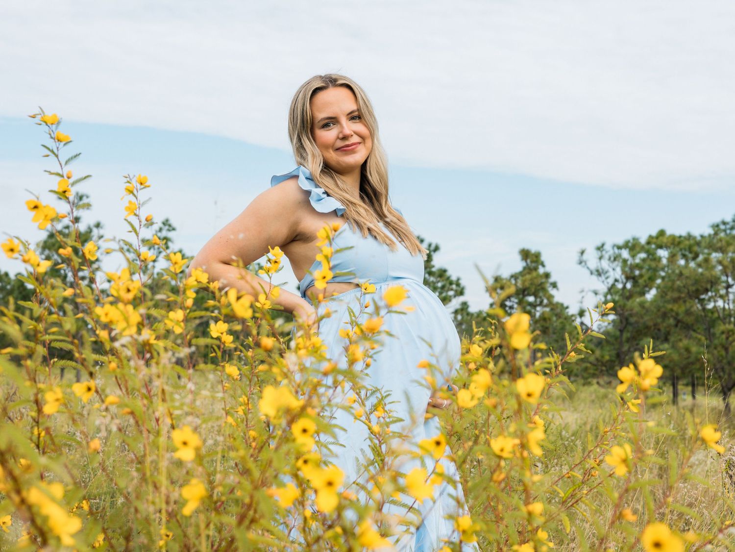 Pregnant woman in blue dress smiling among yellow wildflowers in a sunny field.