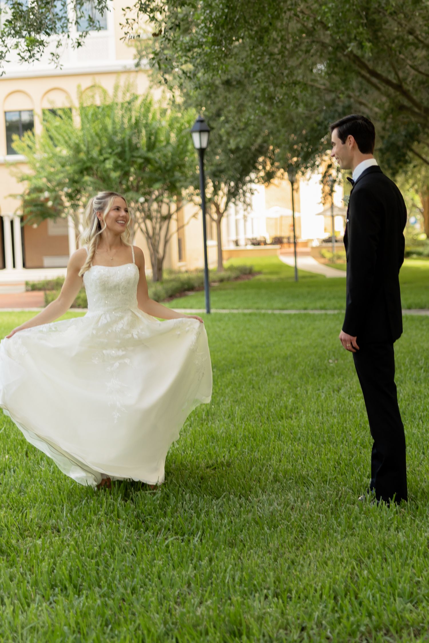 Bride in white gown twirling on green lawn while groom in black tuxedo watches during outdoor wedding photo session.