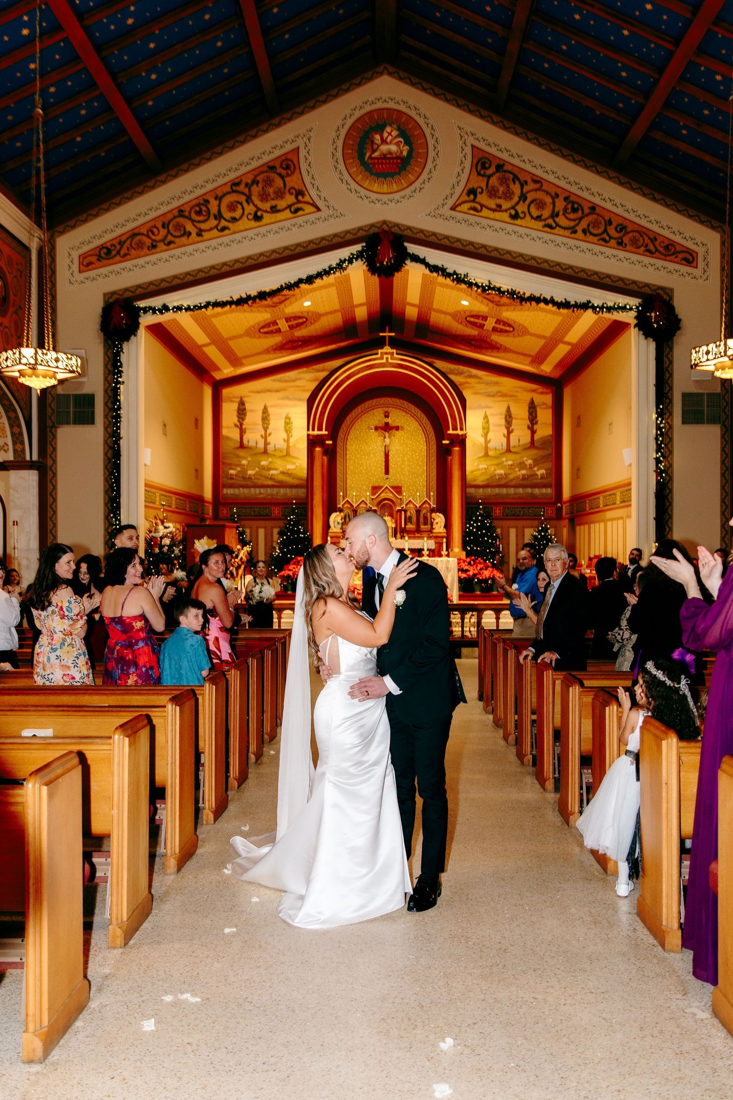 Bride and groom share first kiss in ornate church aisle with Christmas decorations and golden altar backdrop.