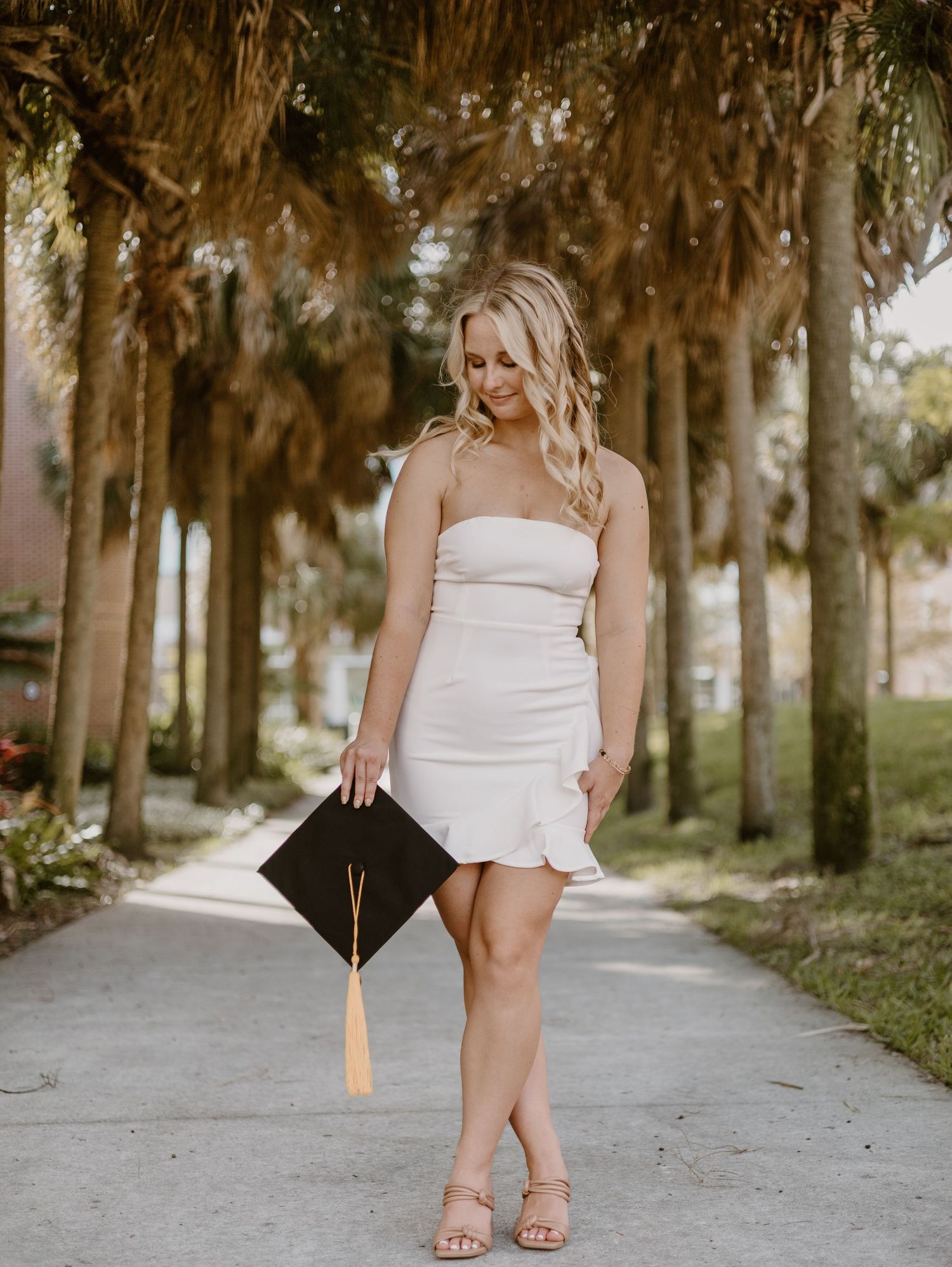 A blonde woman in a white strapless mini dress holds a graduation cap while posing on a palm tree-lined path.