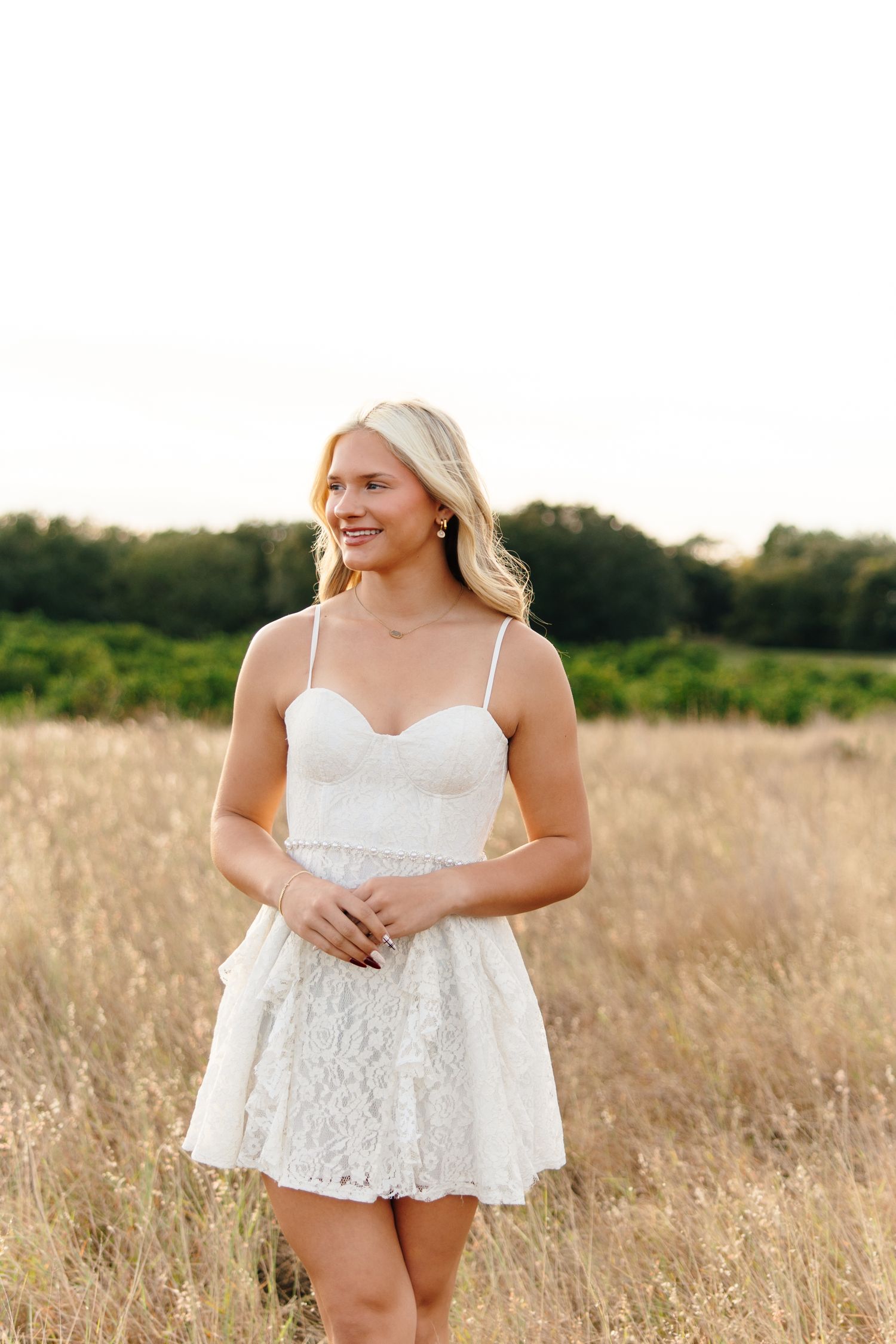 Young blonde woman in white lace mini dress smiling in golden field with green trees in background.
