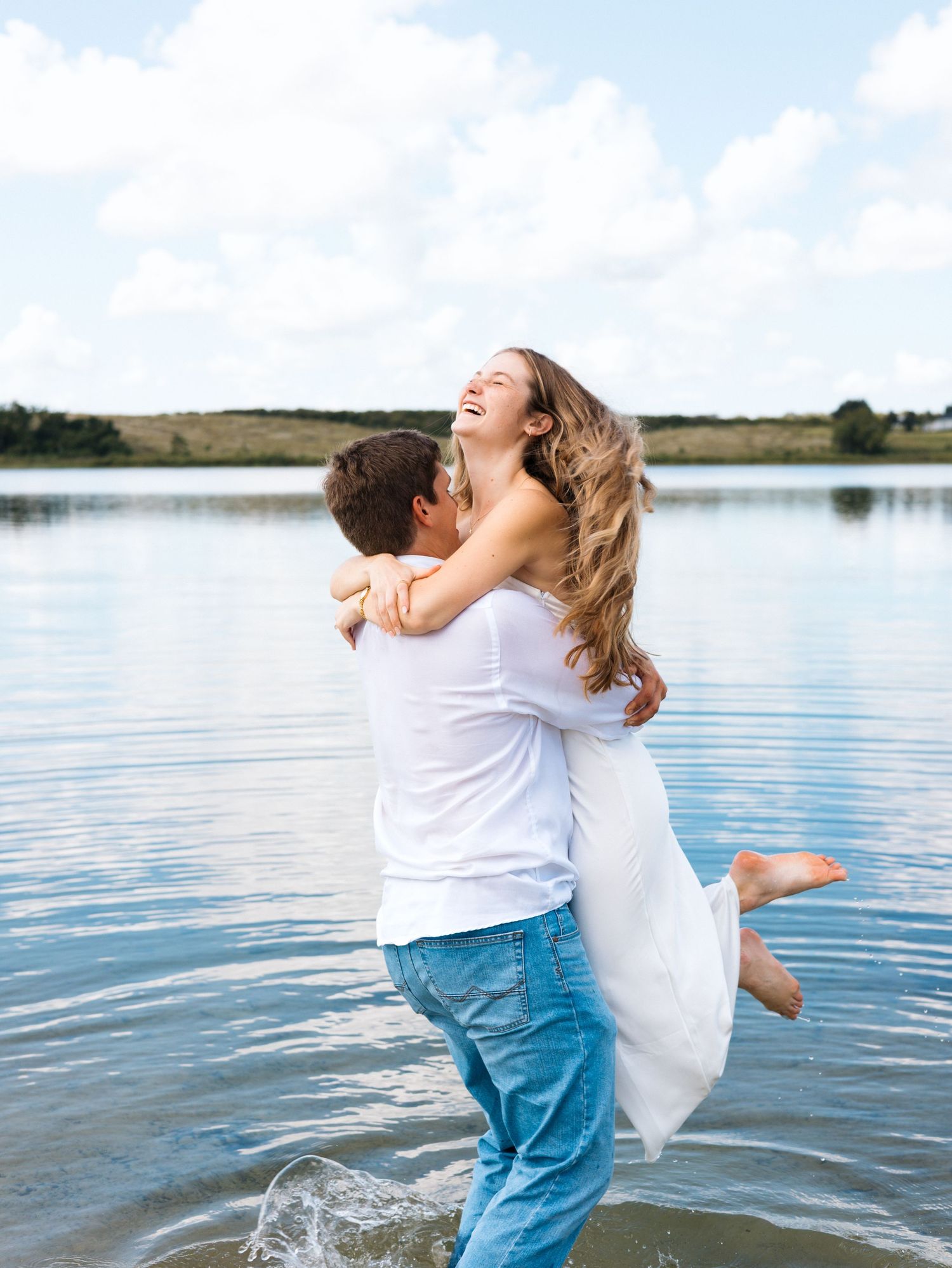 Couple laughing and embracing by a lake, man lifting woman in white dress, joyful romantic outdoor moment.