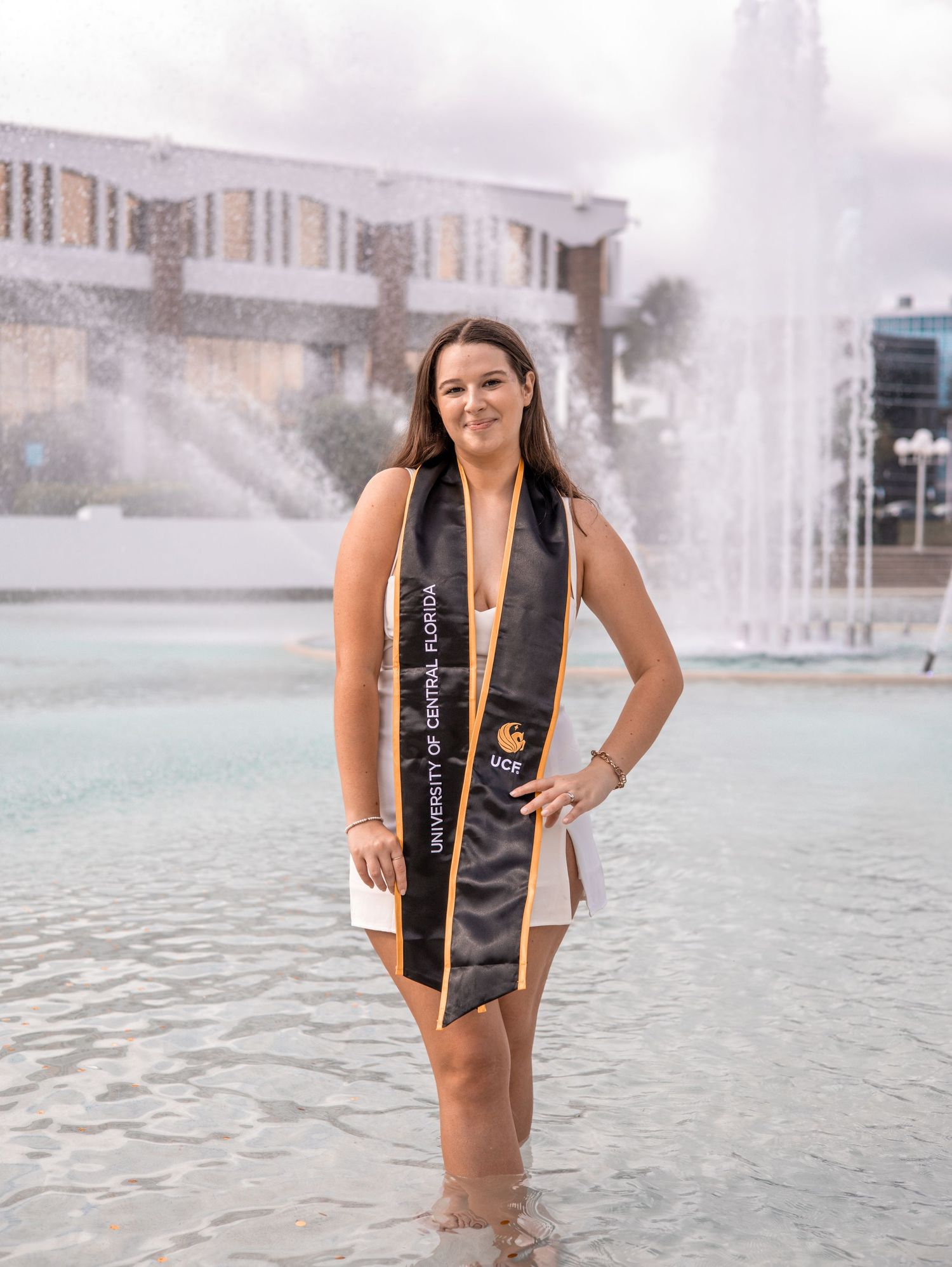 UCF graduate smiles in black and gold stole standing in fountain at University of Central Florida campus.