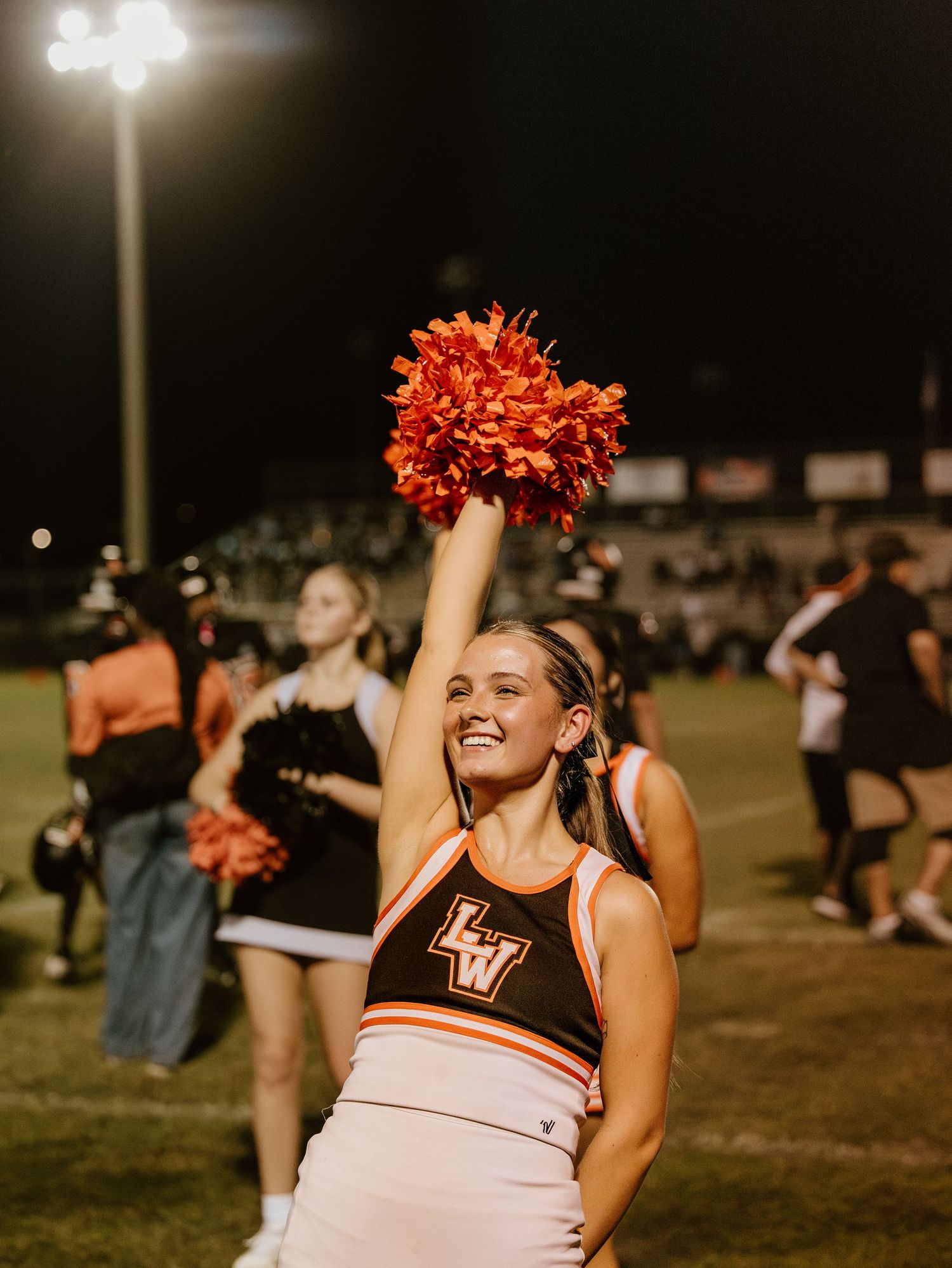 Cheerleader in black uniform with orange pom poms performs energetic routine at nighttime sports event.