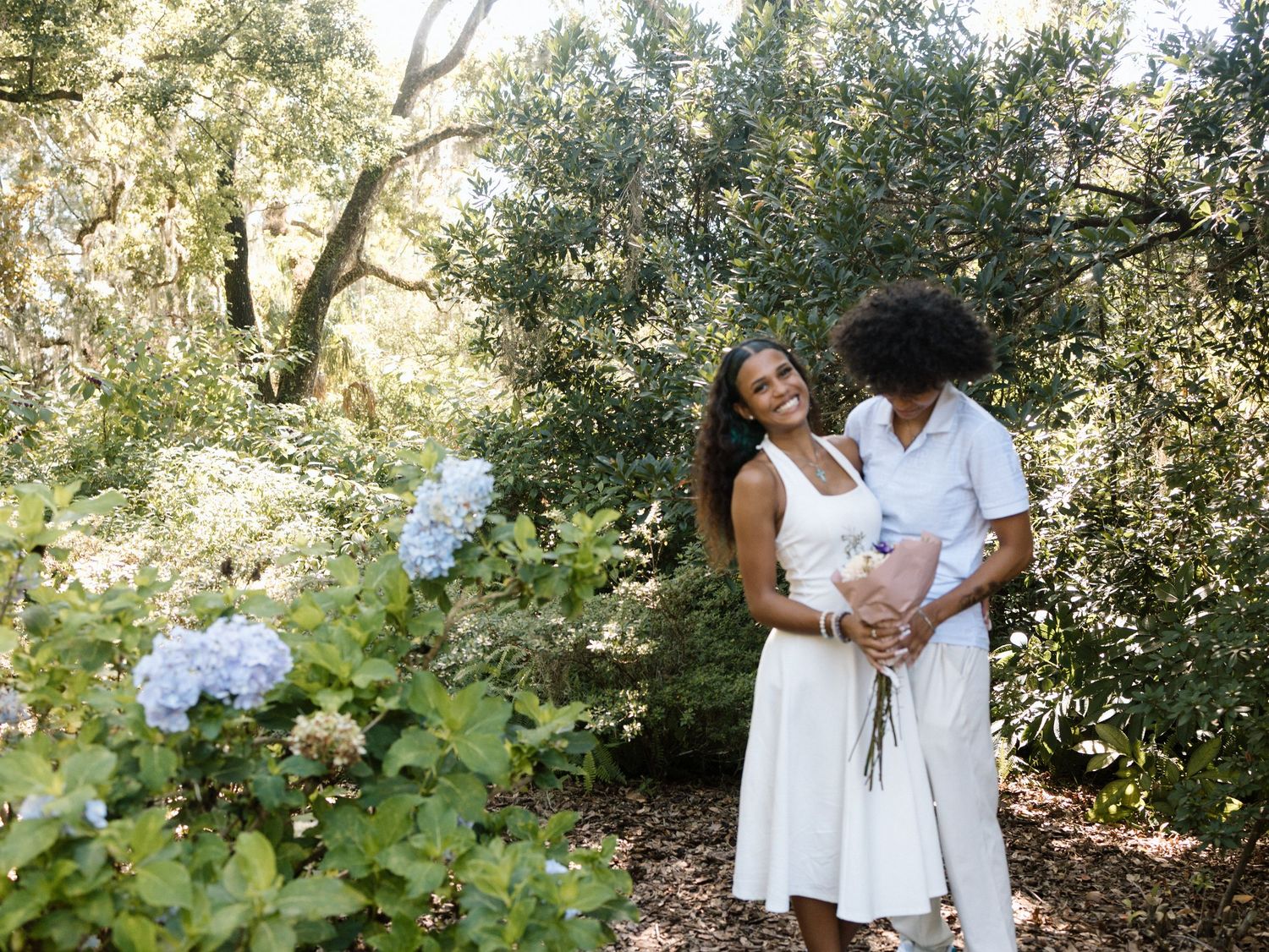 A romantic couple stands together in a garden setting with blooming hydrangeas and dappled sunlight filtering through trees.