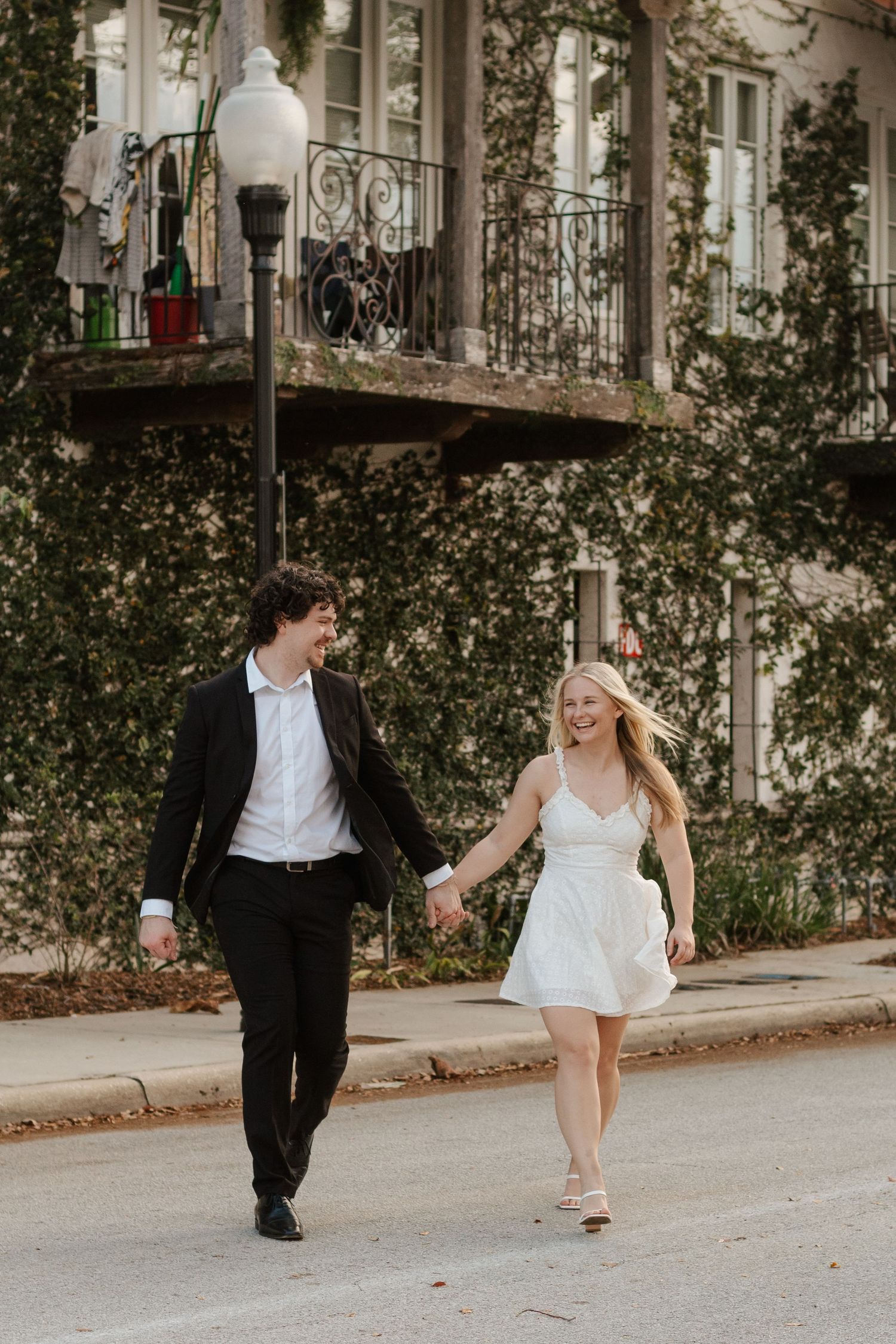 A couple runs playfully along a tree-lined street in formal attire.