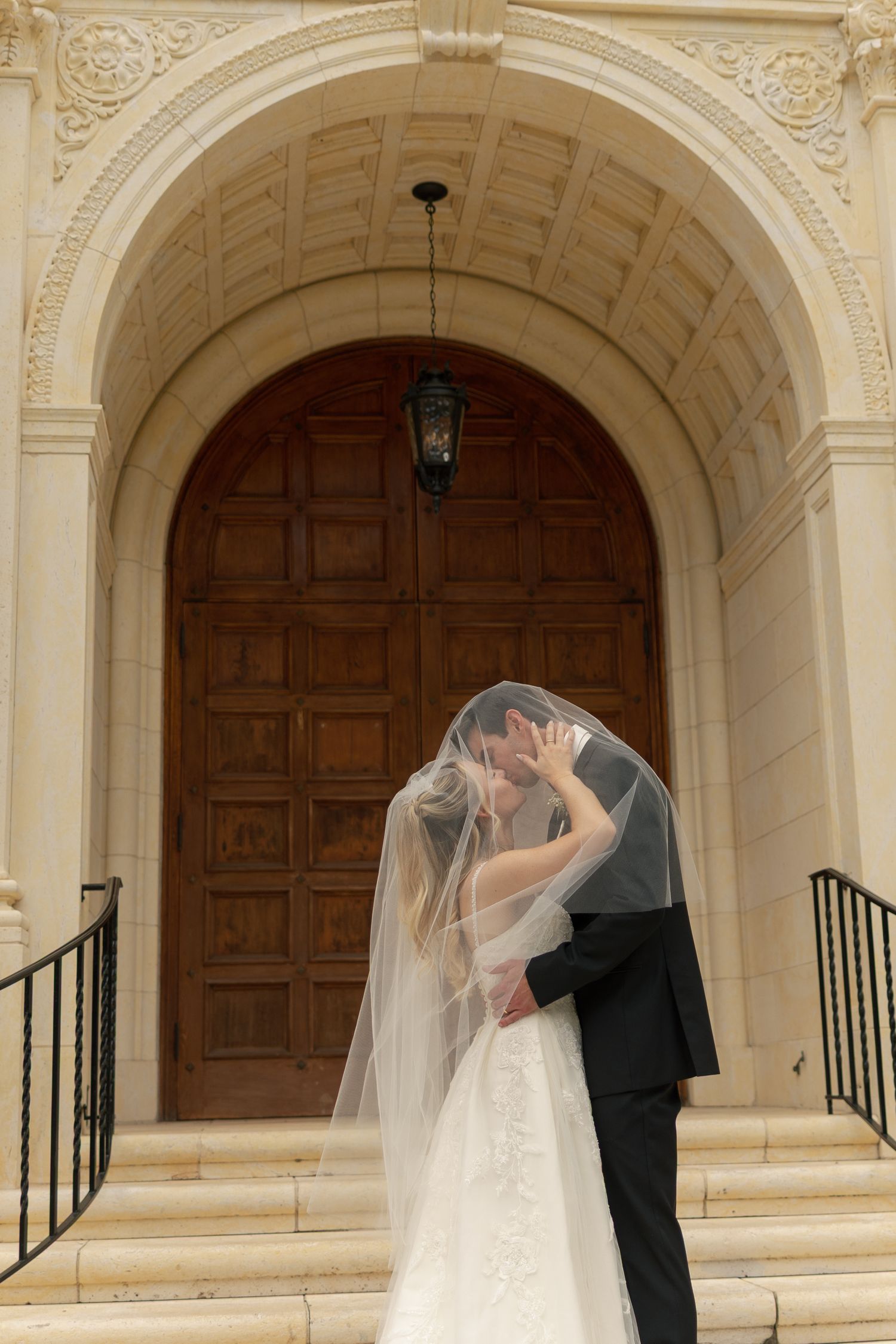 Bride and groom sharing a kiss under a flowing veil on church steps with ornate wooden doors.
