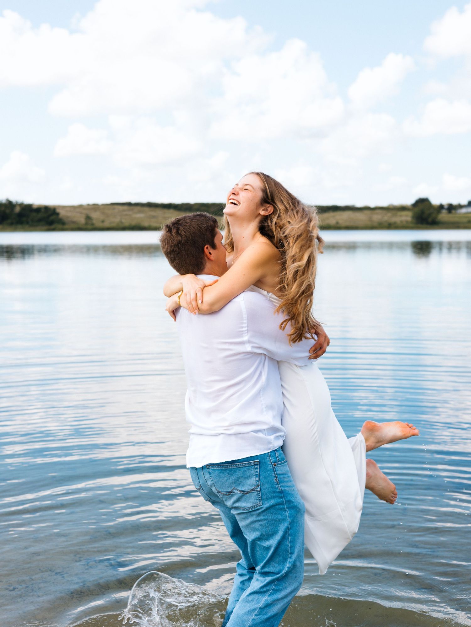 A couple in white shirts and jeans embraces playfully by a serene lake on a sunny summer day.