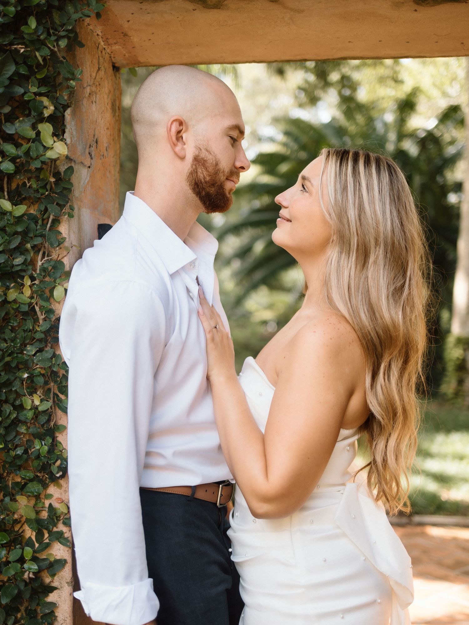 A couple in white wedding attire share a romantic moment under a rustic ivy-covered archway outdoors.