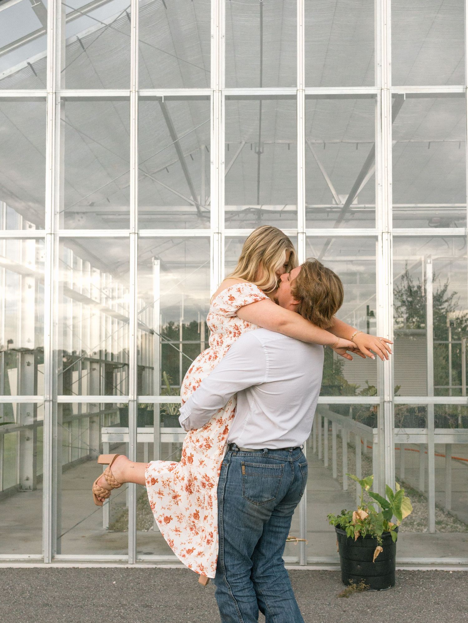 A couple shares a romantic embrace in front of a glass greenhouse wall during a photo session.