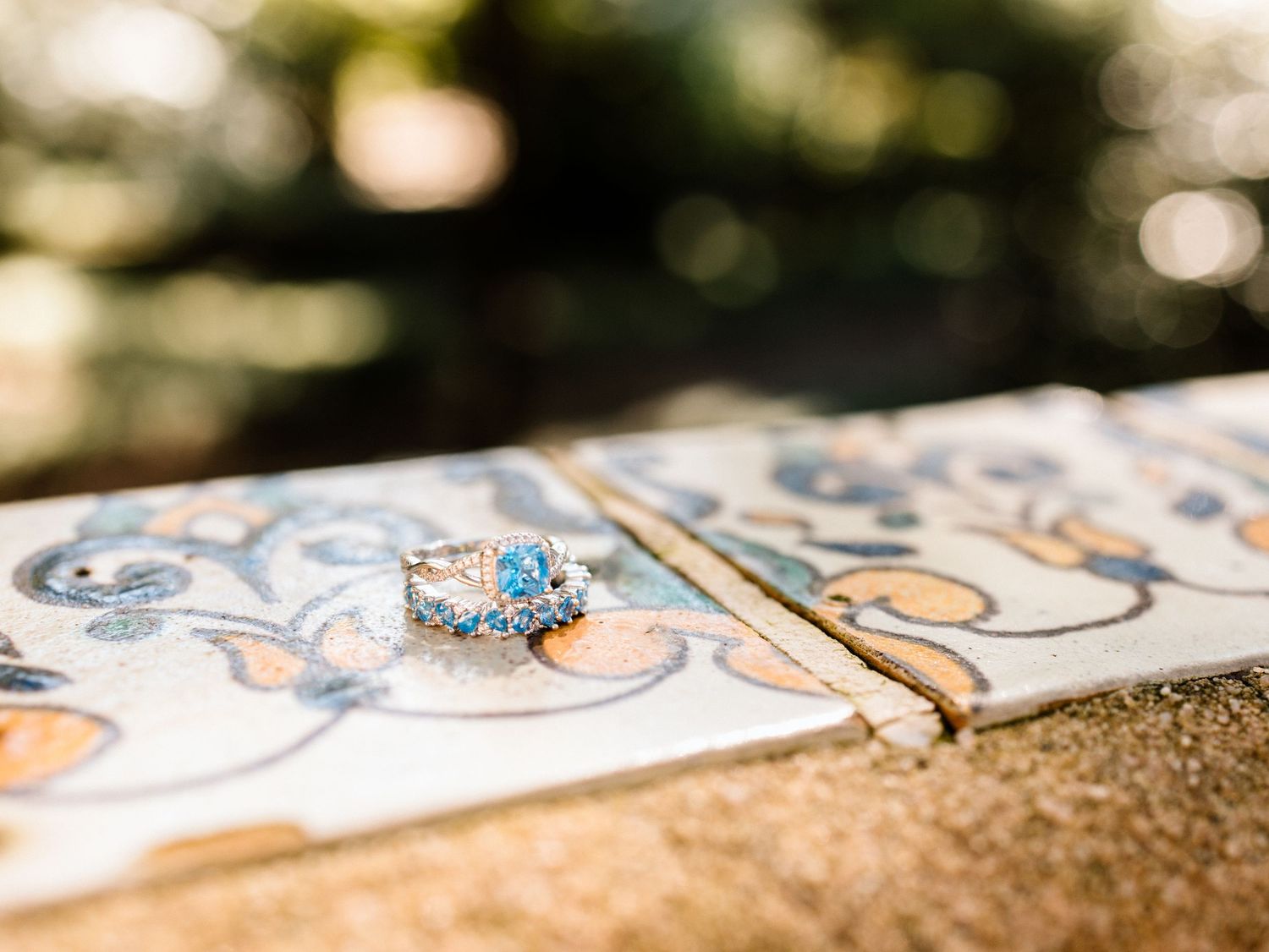 Delicate blue engagement ring photographed on decorative tile with soft bokeh background.