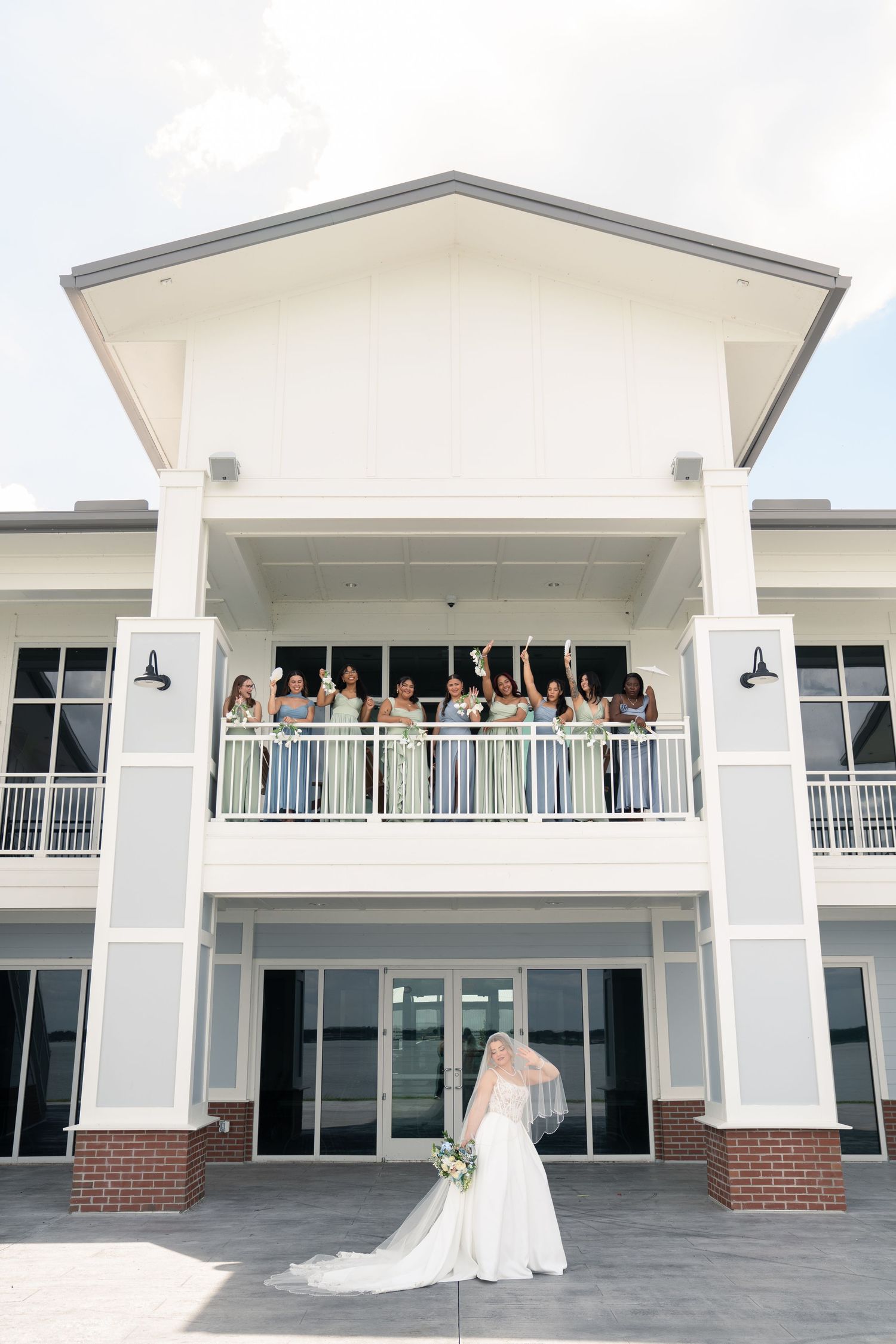 Bride in white gown stands before venue as bridesmaids cheer from balcony above on wedding day.