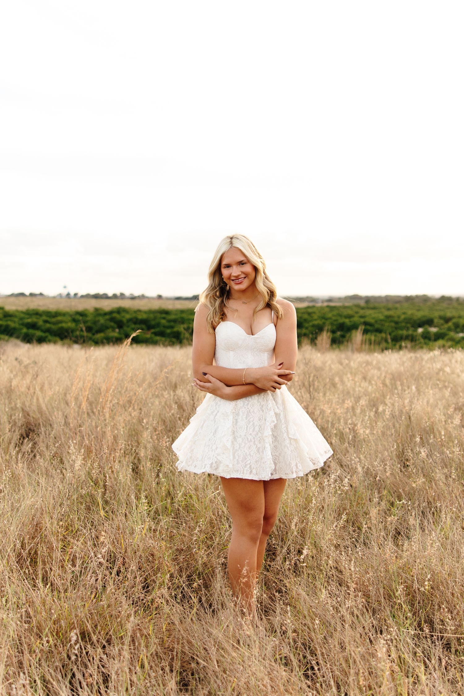 Smiling blonde woman in white strapless mini dress standing in golden field with green trees in background.