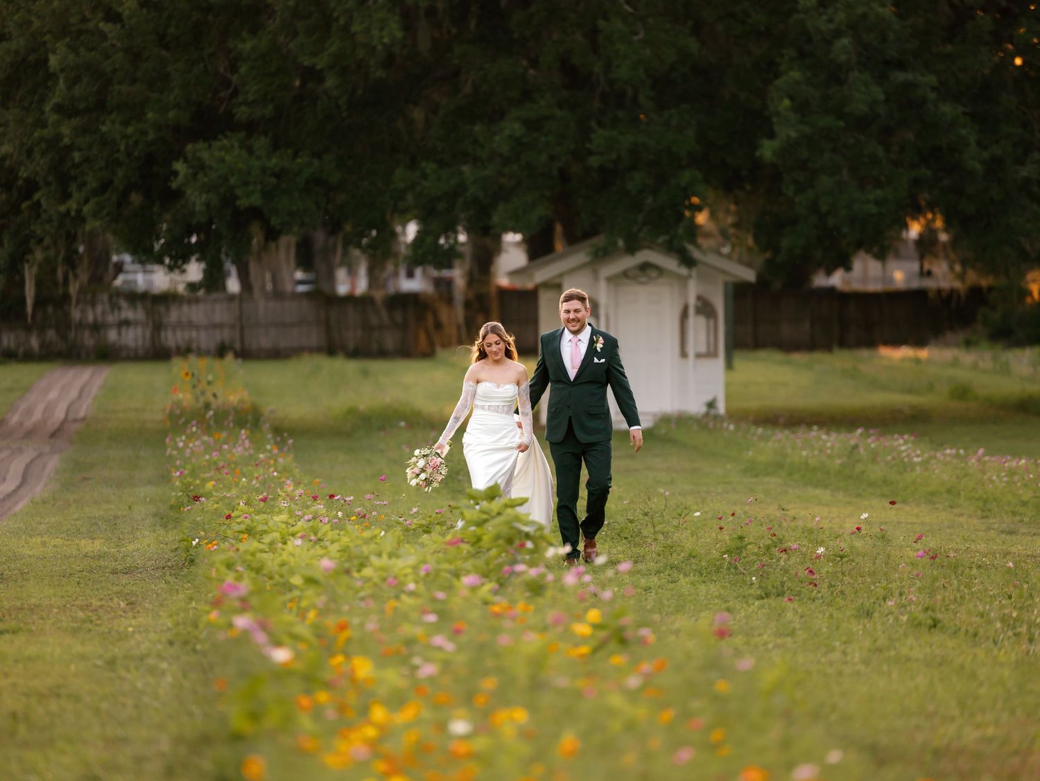 Bride and groom walk hand-in-hand through a lush green meadow with wildflowers on their wedding day.