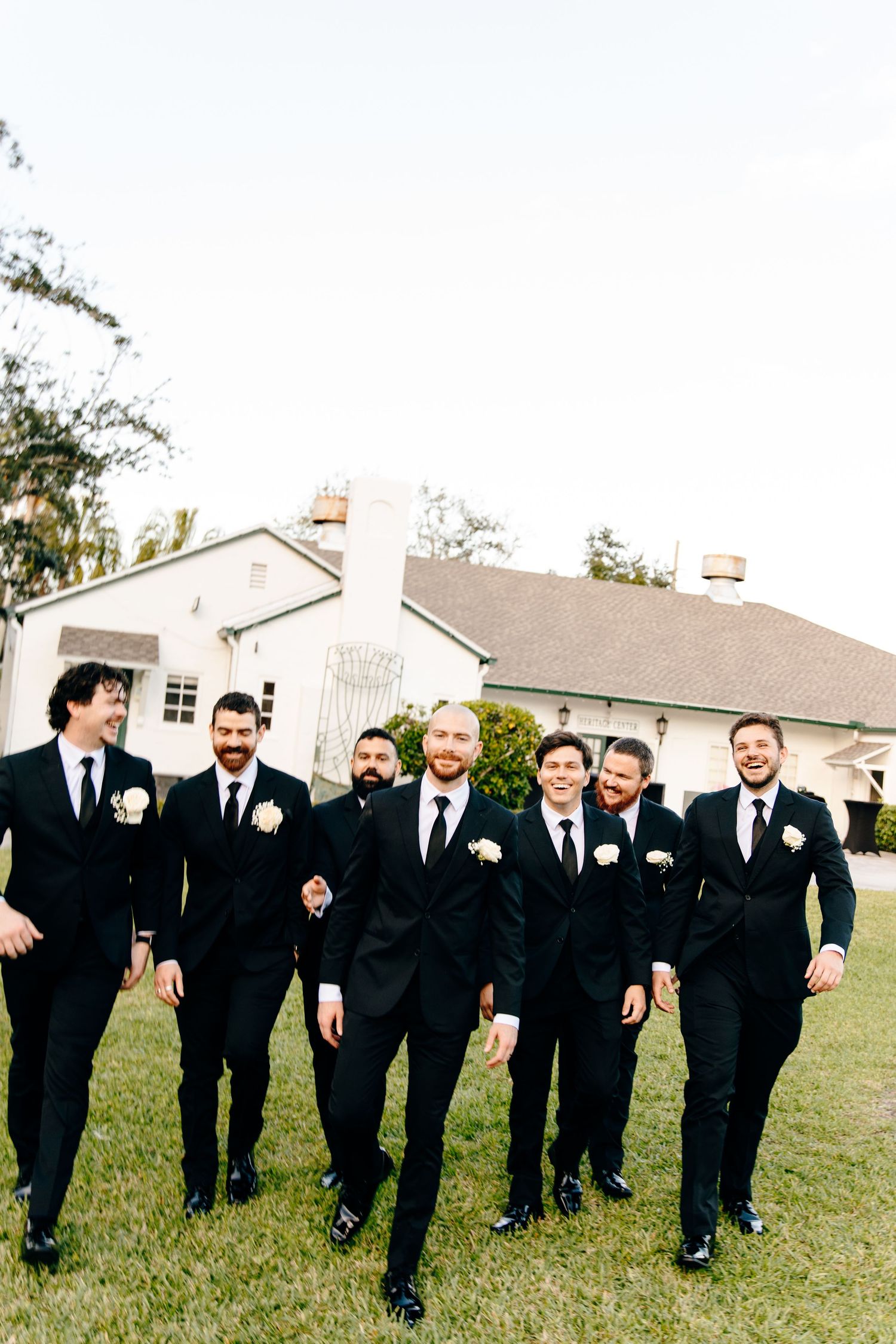 Groom and groomsmen in black suits with white boutonnieres walk confidently across a green lawn together.