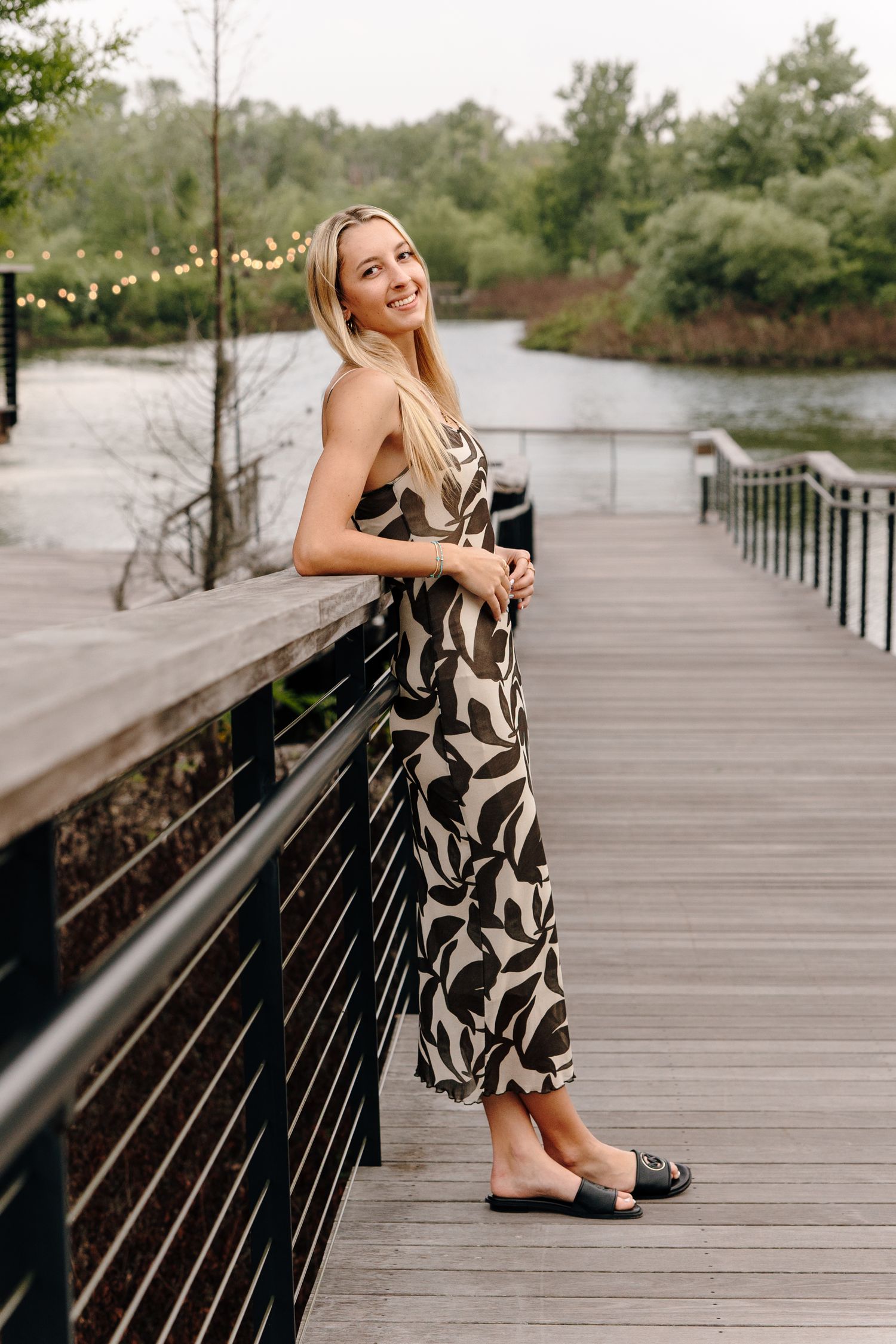 Smiling blonde woman in a floral jumpsuit posing on a wooden boardwalk by a serene river surrounded by lush greenery.