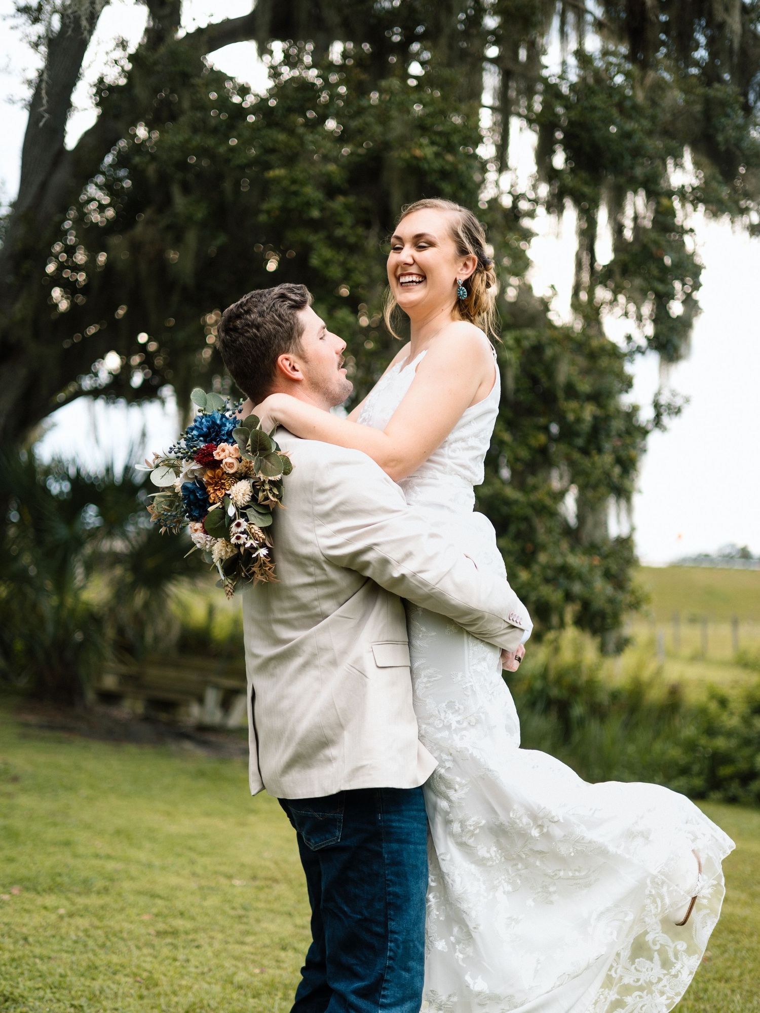 A joyful wedding couple embraces outdoors under mossy trees, with the bride in white lace and groom in light blazer.