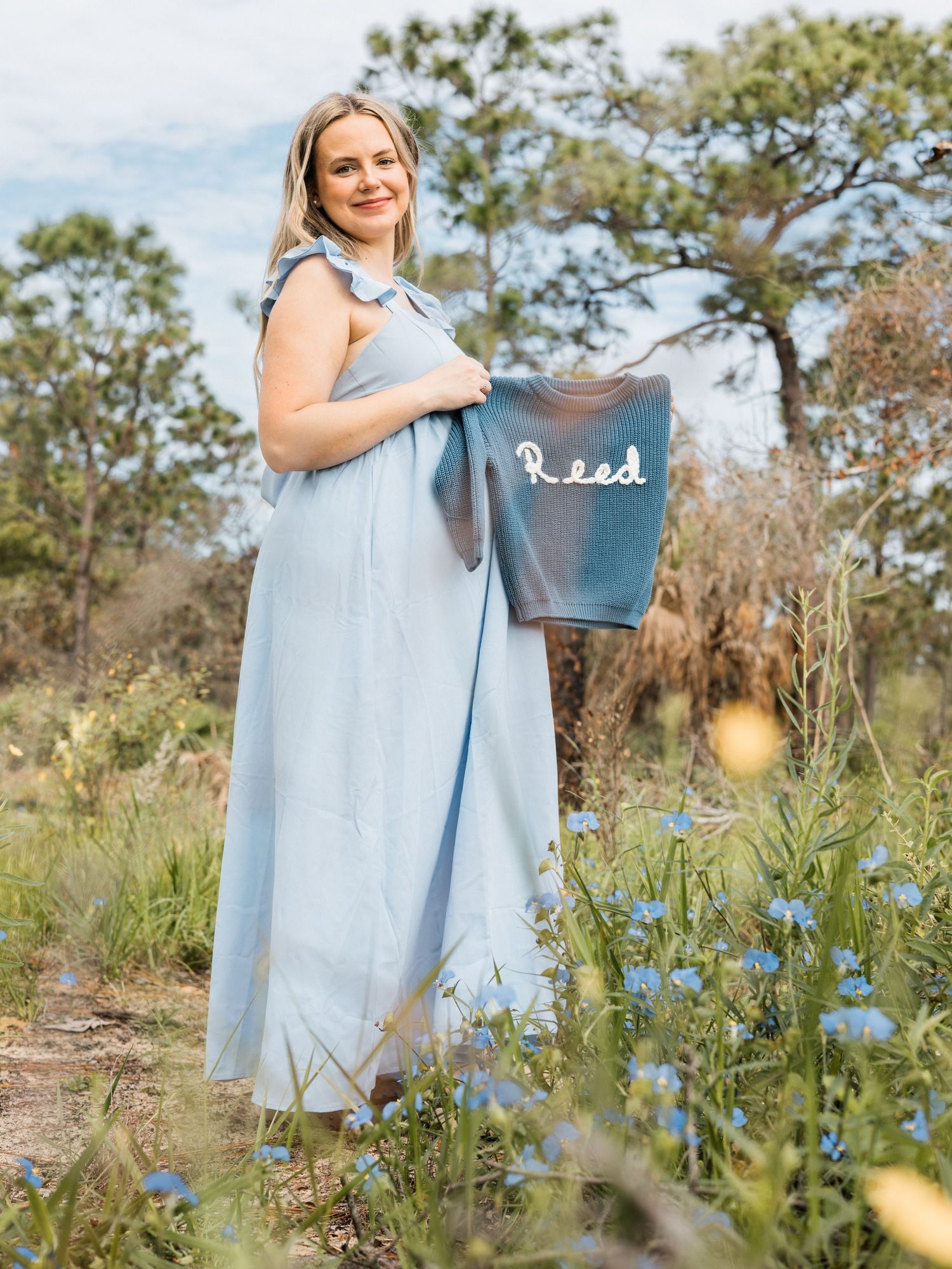 Figure in light blue dress stands in sunny wildflower field holding watering can against natural outdoor backdrop.