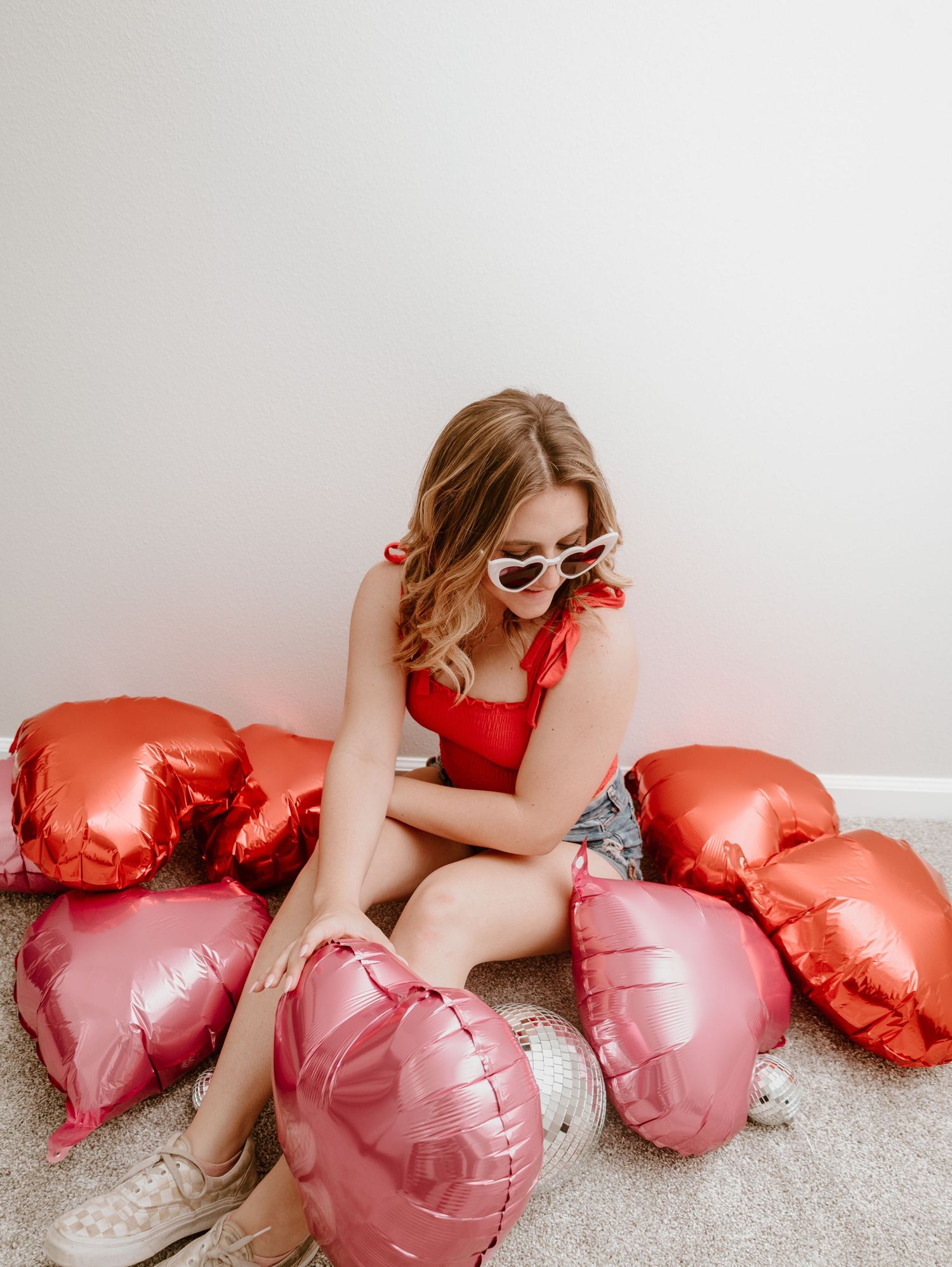 A person reclines with heart-shaped mylar balloons in pink and red colors on a light gray background.