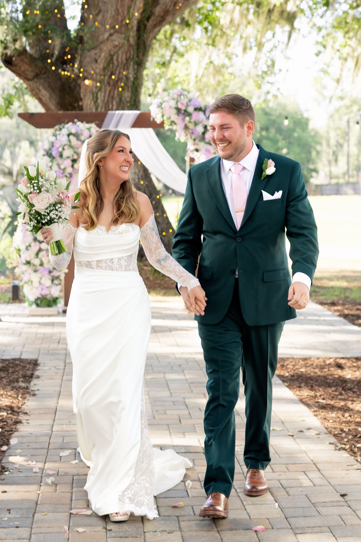 Smiling bride and groom walk hand-in-hand on brick path, she in white lace gown, he in dark green suit with pink tie.