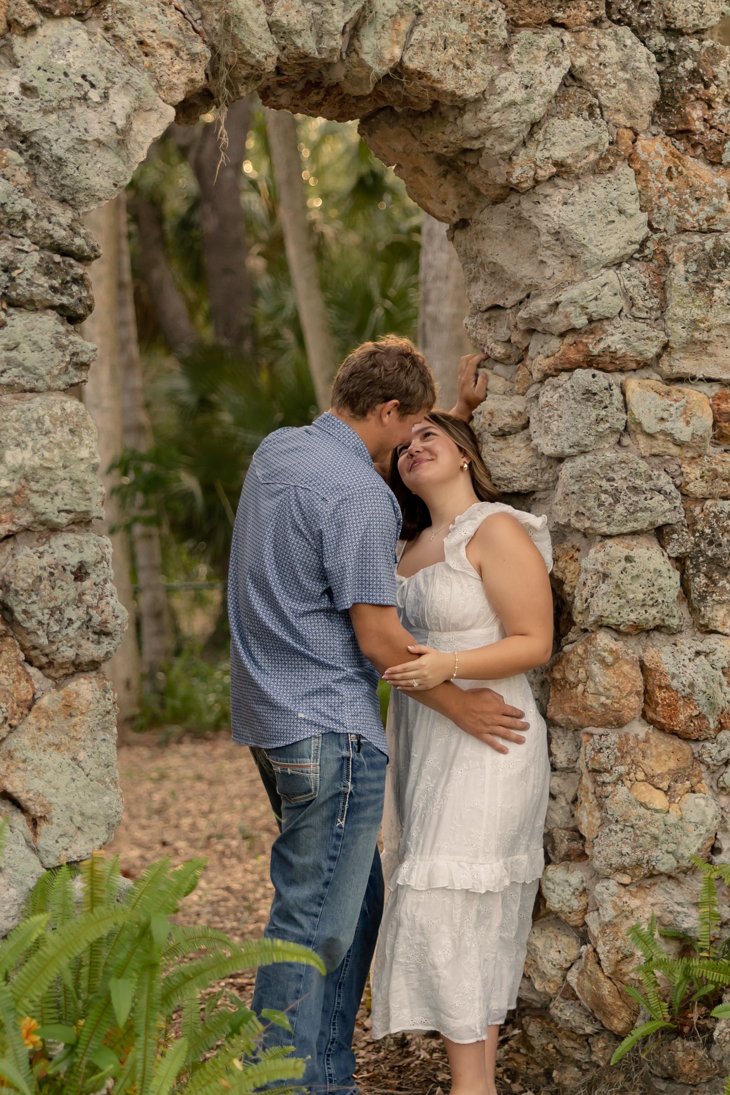 Couple sharing a romantic moment against rustic stone arch wall, woman in white dress smiling up at man in blue shirt.