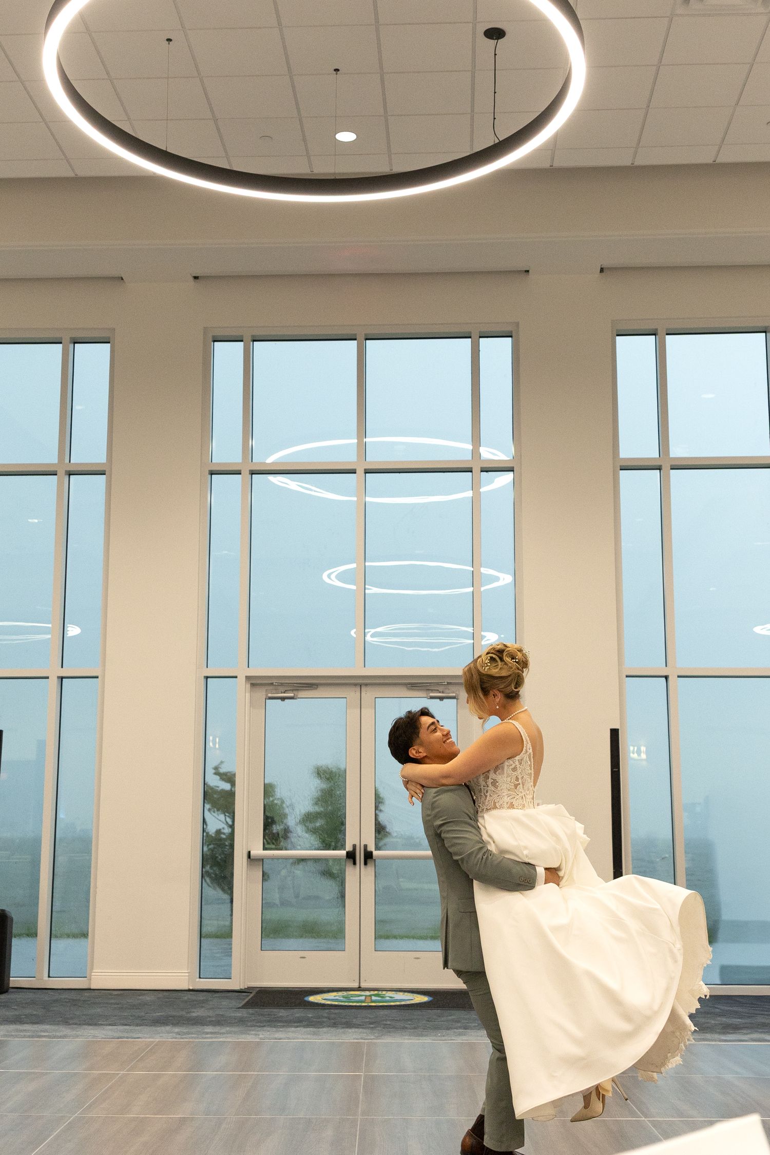 Groom lifts bride in white wedding dress during first dance in elegant venue with large windows.