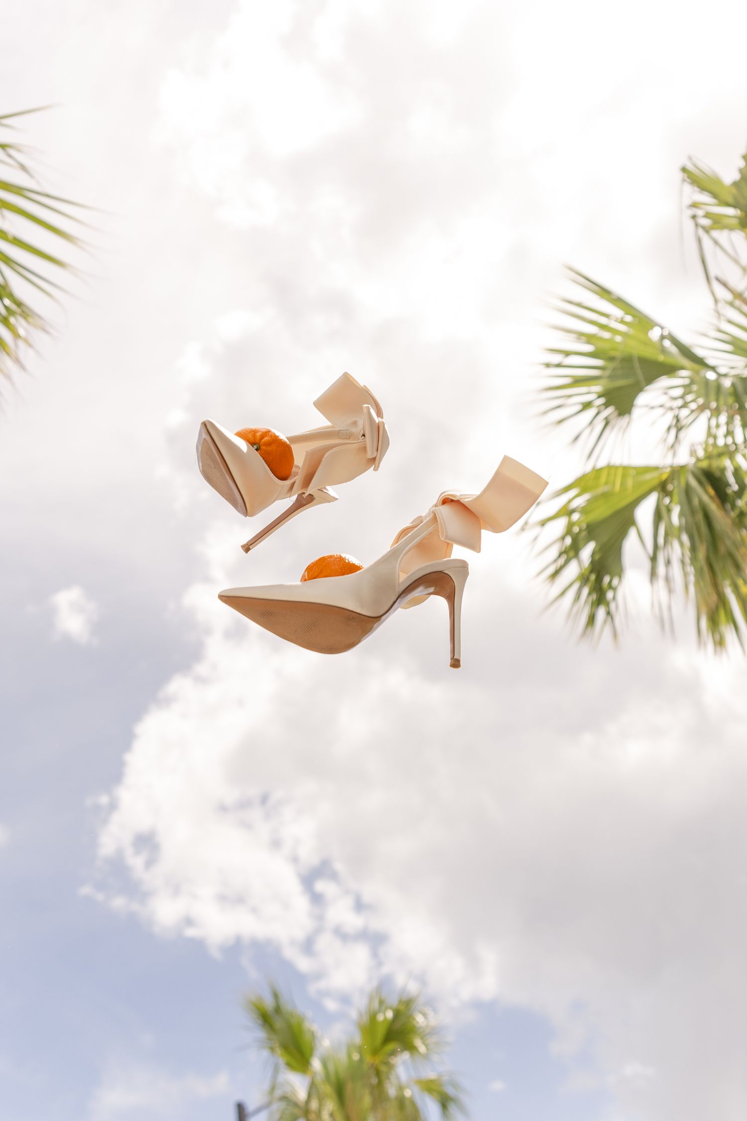 Elegant cream bridal heels with satin bows tossed in the air against a cloudy sky framed by tropical palm trees.