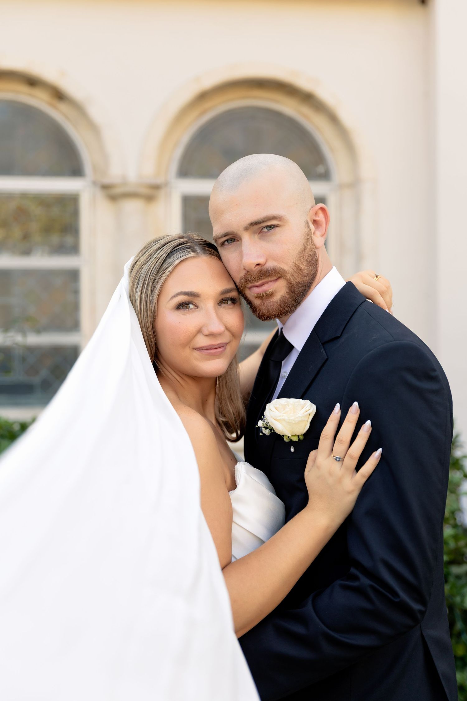A bride and groom embrace tenderly outdoors, she in a white gown with veil, he in a dark suit with boutonniere.