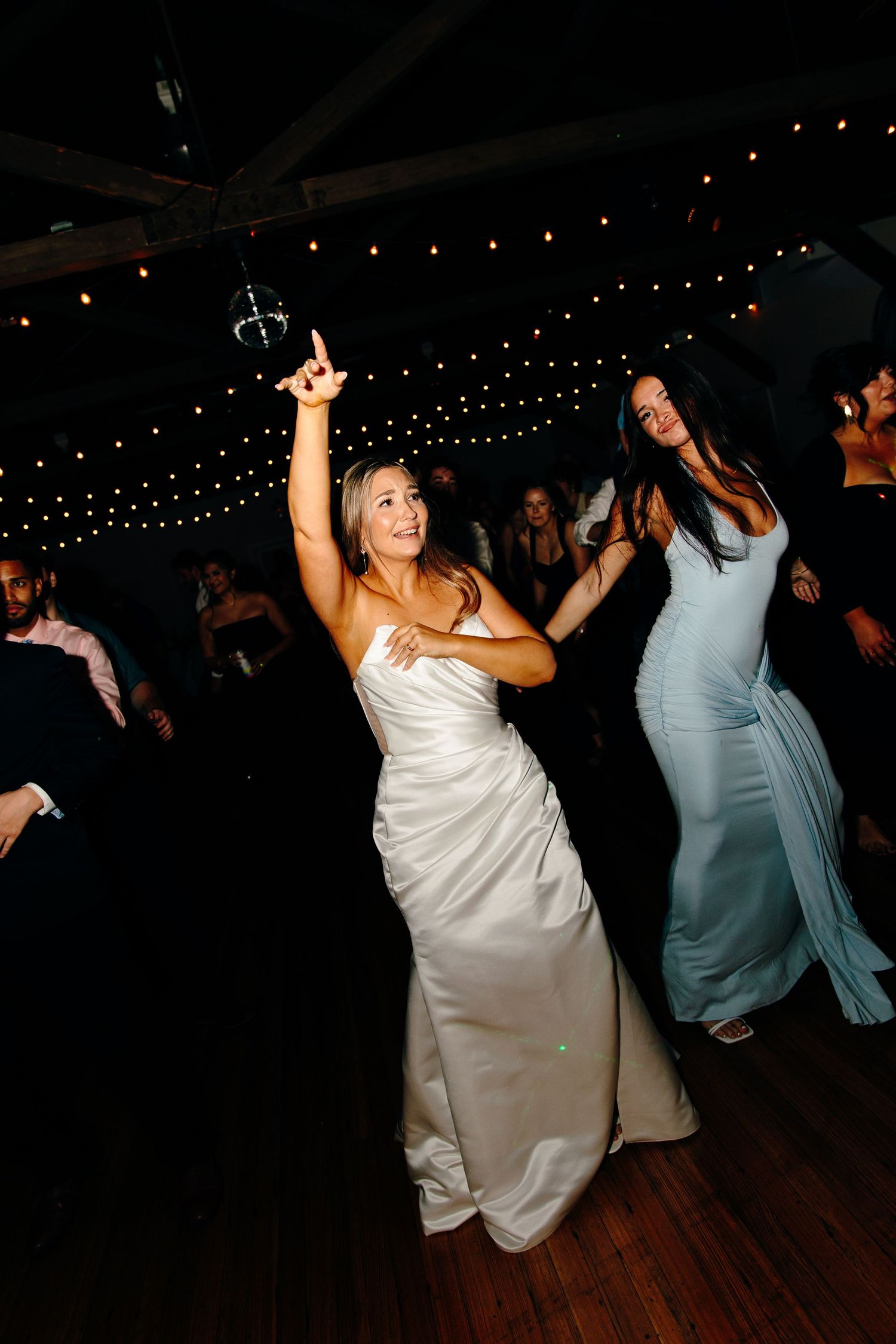 Bride in white gown dancing joyfully at wedding reception with guests and string lights in background.