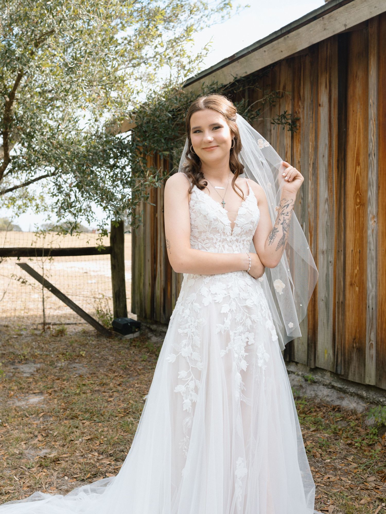A bride in a flowing white lace wedding dress poses in front of a rustic wooden barn building during a photo session.