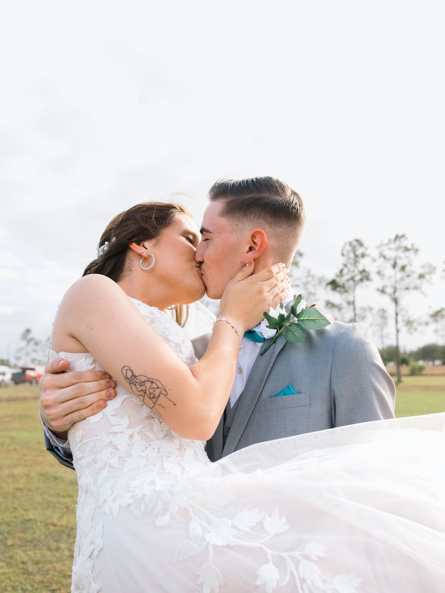A romantic couple shares tender kisses while embracing outdoors in a field during their wedding photoshoot.
