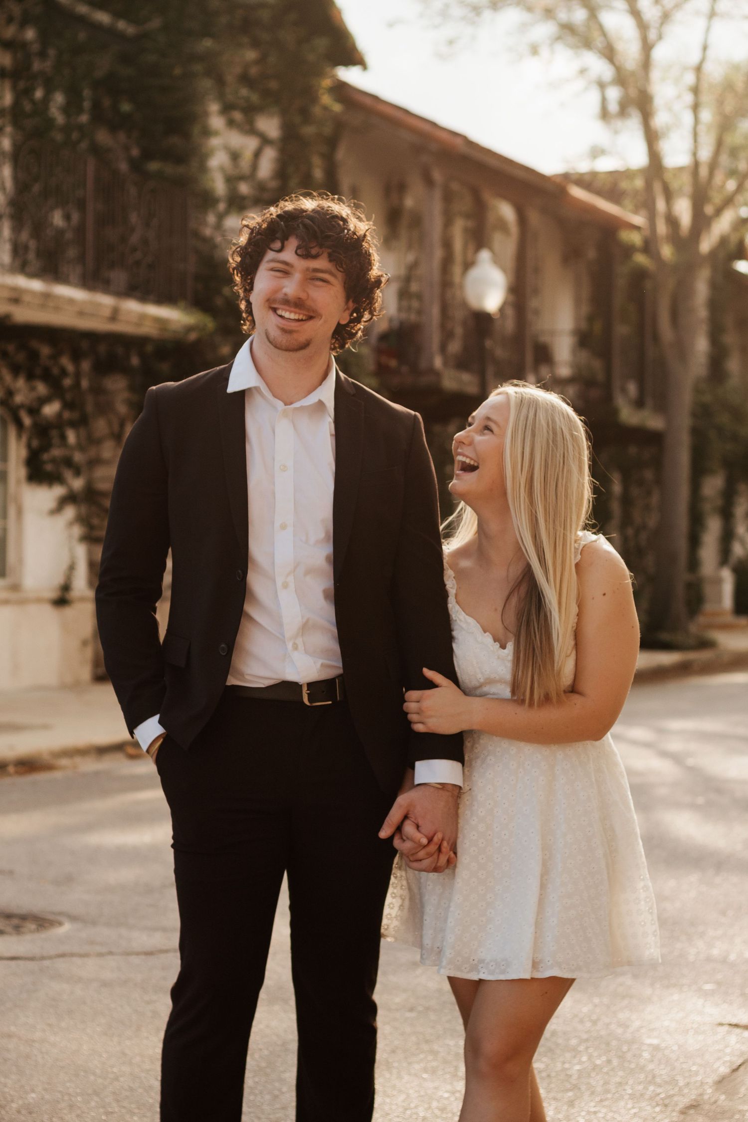 A couple in formal attire shares candid laughter and affection in a sun-dappled alleyway.