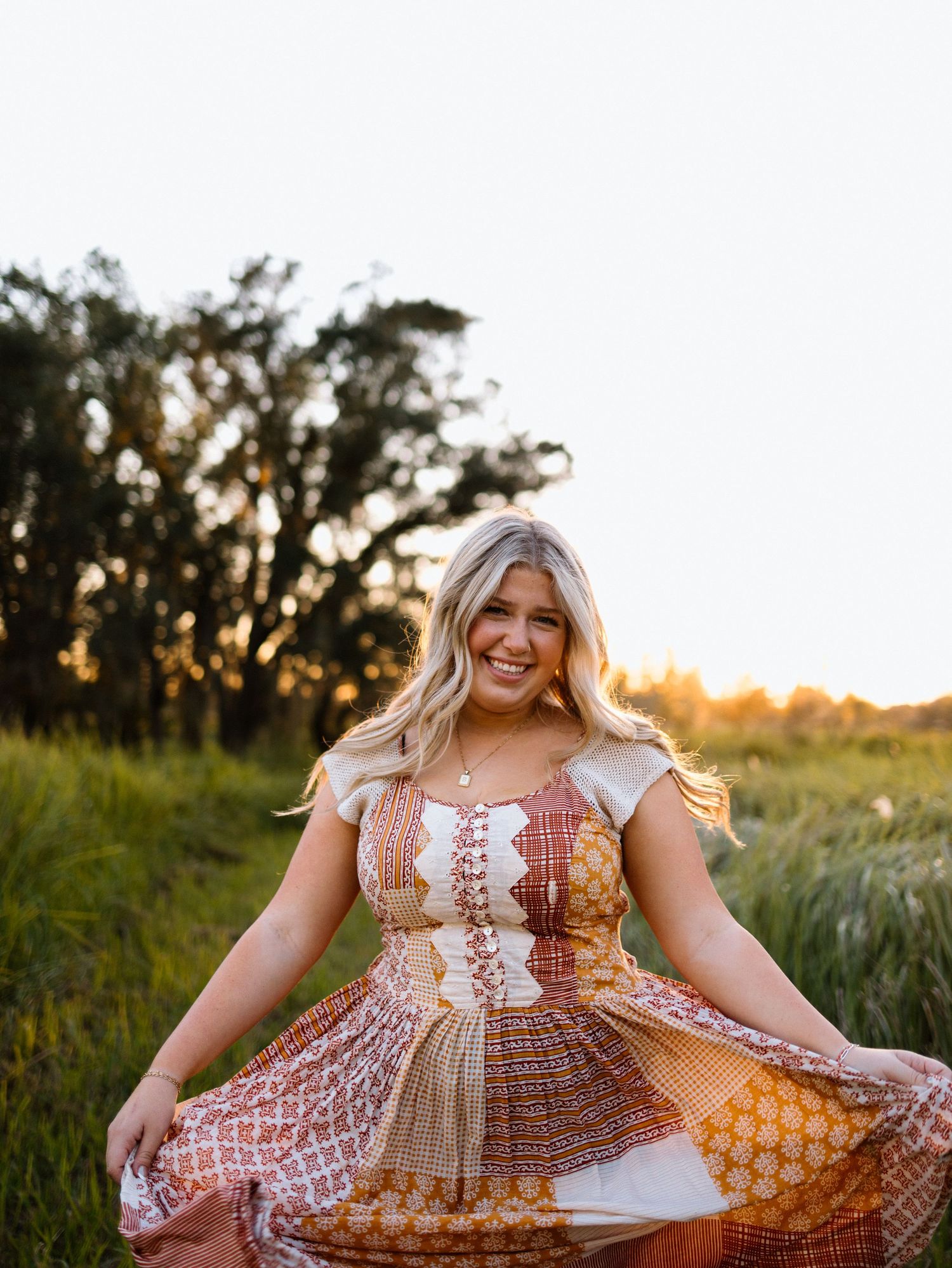 Woman in prairie dress spreads skirt while standing in golden sunset field amidst tall grass.